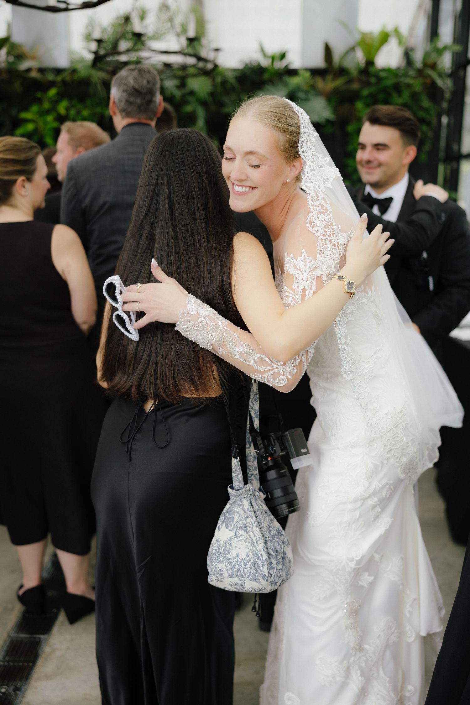 Smiling bride in lace wedding gown and veil embraces a guest during outdoor wedding reception.