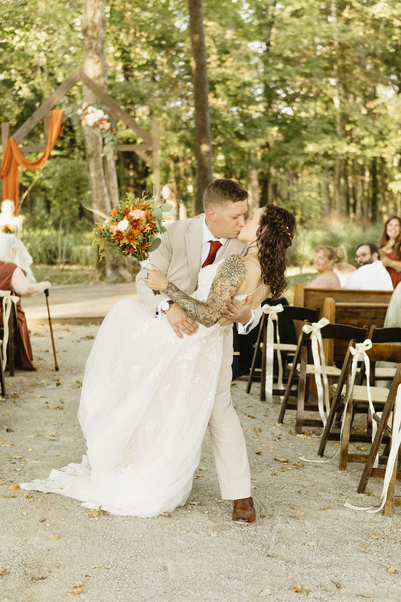Romantic wedding couple dips and kisses during outdoor reception with rustic wooden seating in wooded setting.