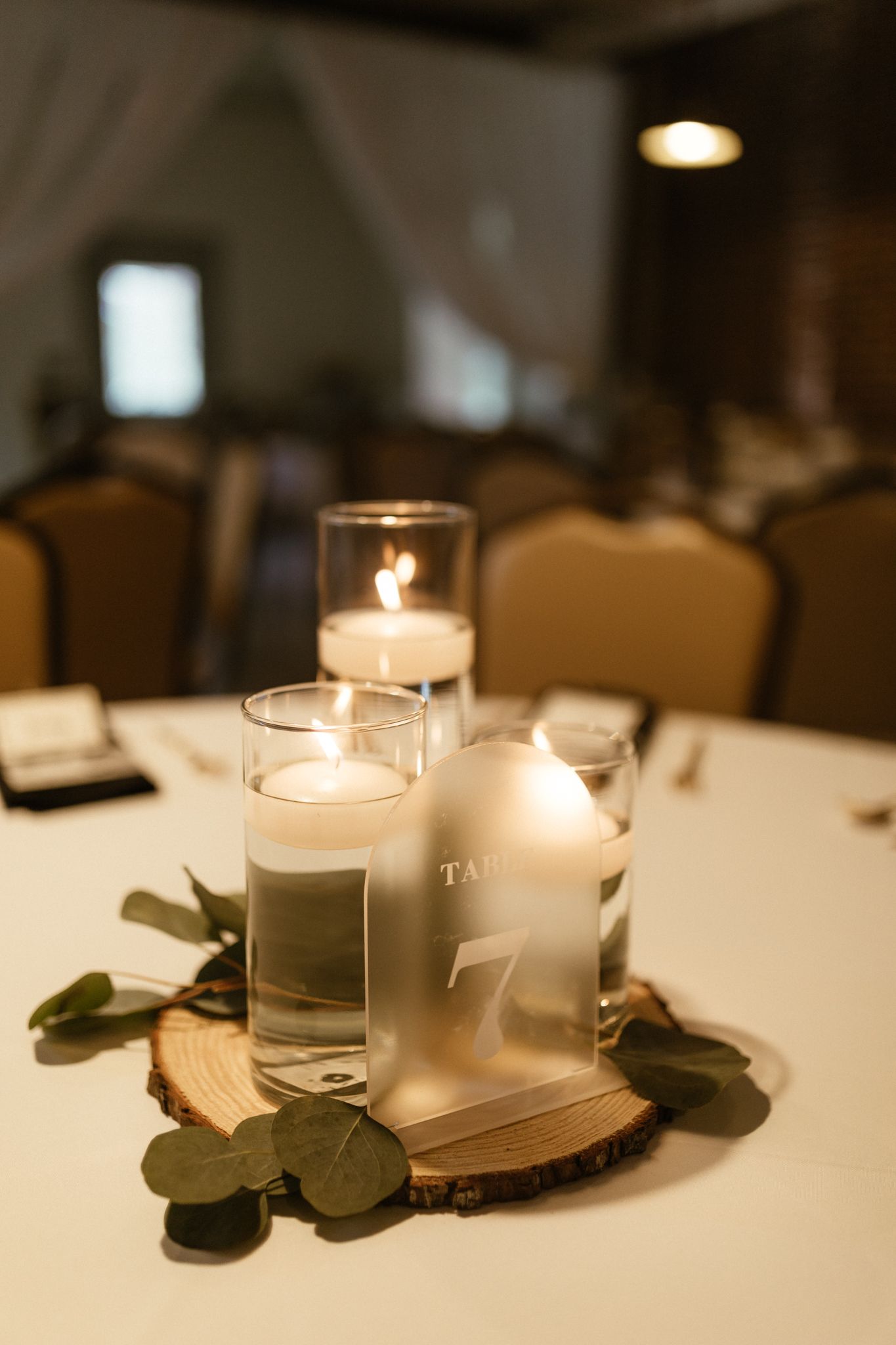 Elegant wedding centerpiece with floating candles in glass cylinders on wood slice with eucalyptus leaves and frosted bottle decor.