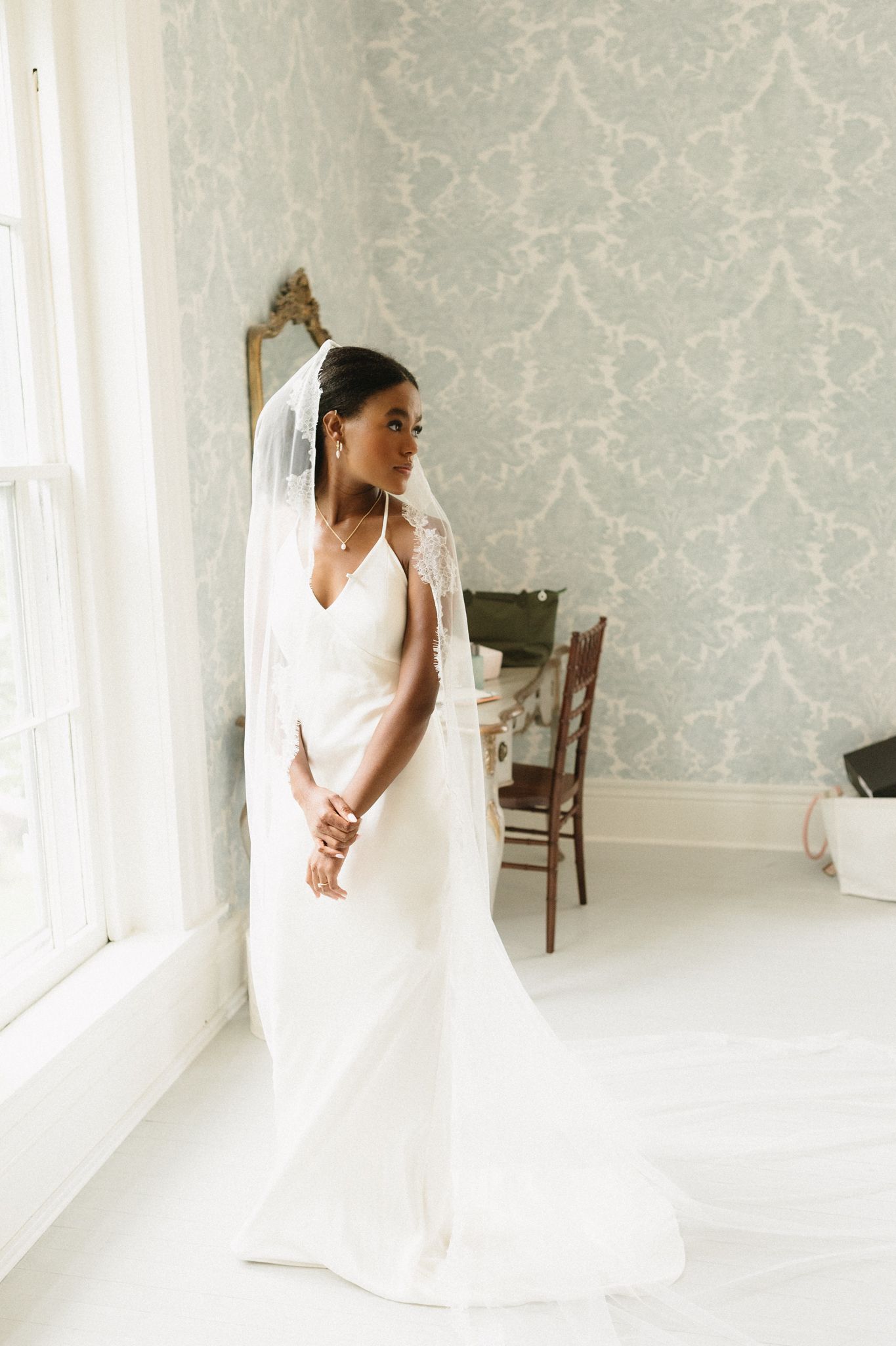 Elegant white wedding dress with long flowing train captured in a bright room with pale damask wallpaper.