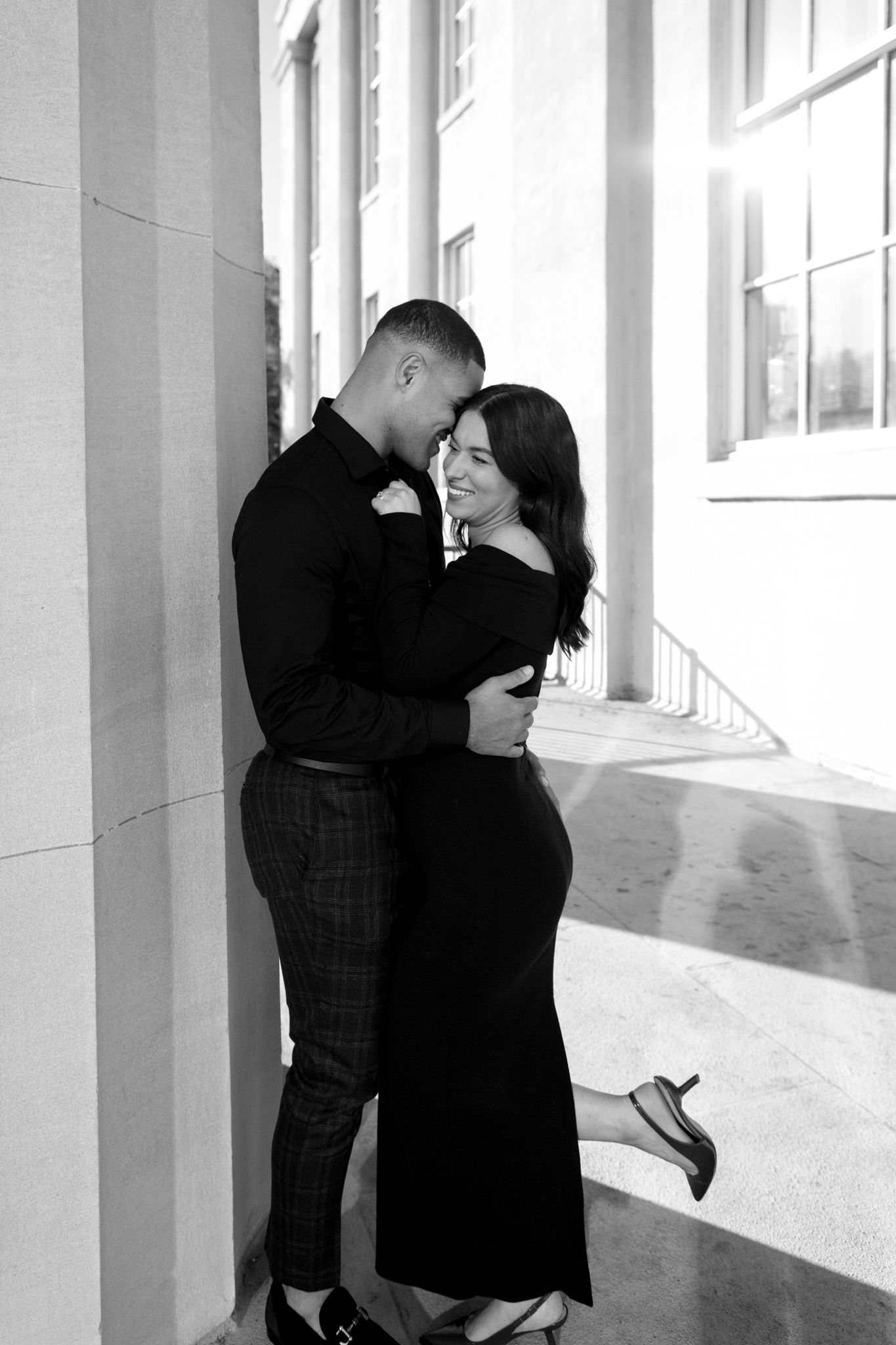 A romantic black and white photo of a couple embracing in front of a bright window with sunlight streaming in.