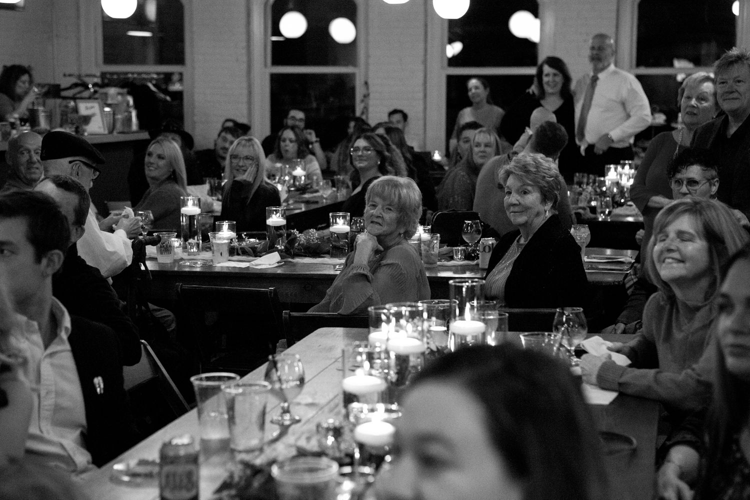 A black and white photograph of diners gathered at tables in a restaurant with candles and glassware.