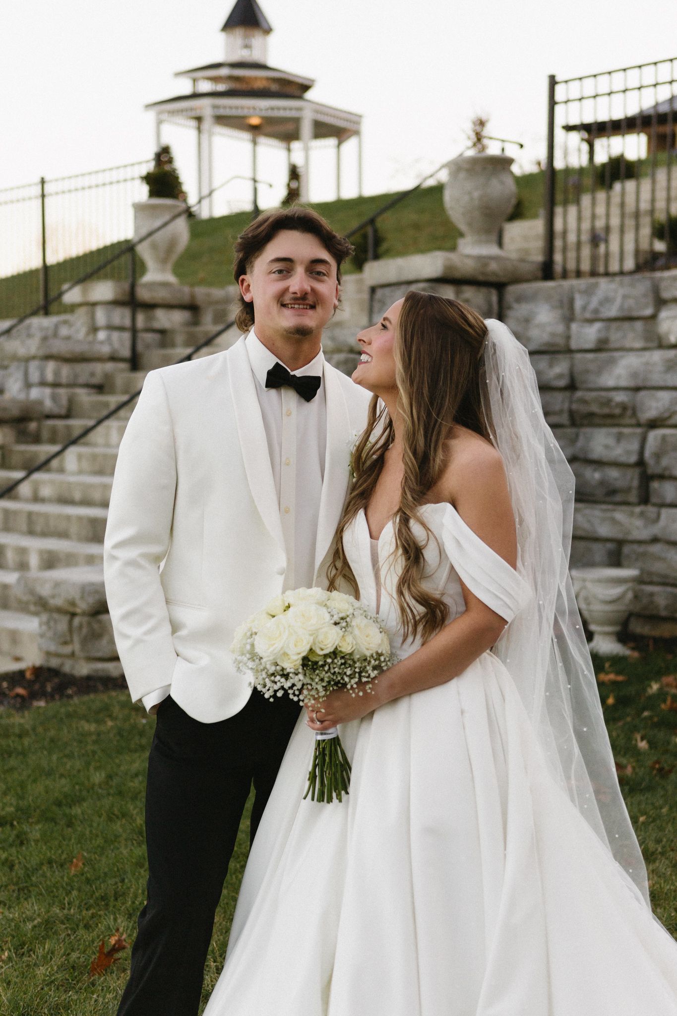 Wedding couple in white attire share intimate moments against stone wall backdrop during outdoor celebration.
