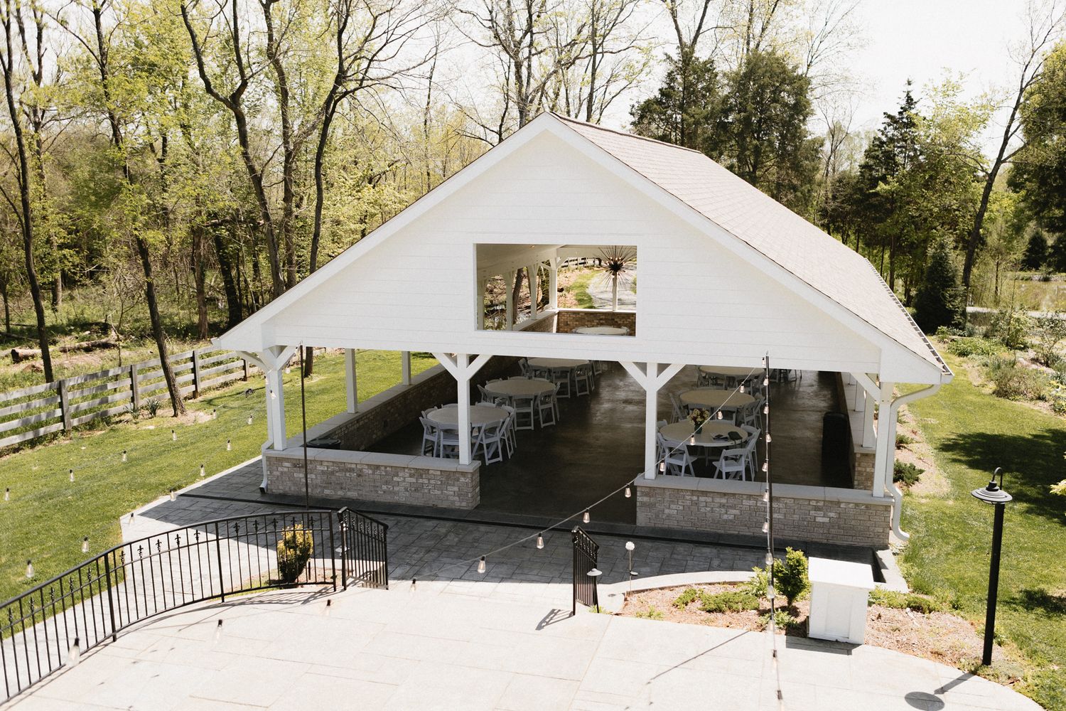 A white outdoor event pavilion with metal railings sits on a concrete pad surrounded by green grass and trees.