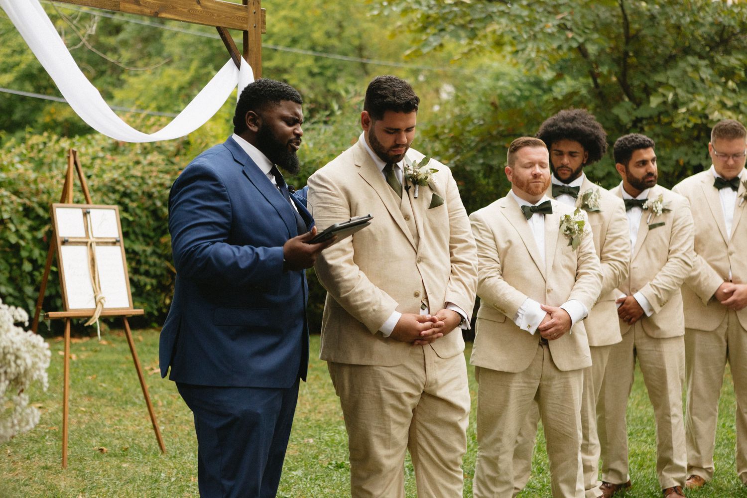 Wedding party outdoors with groomsmen in beige suits and one in navy blue standing at outdoor ceremony altar.