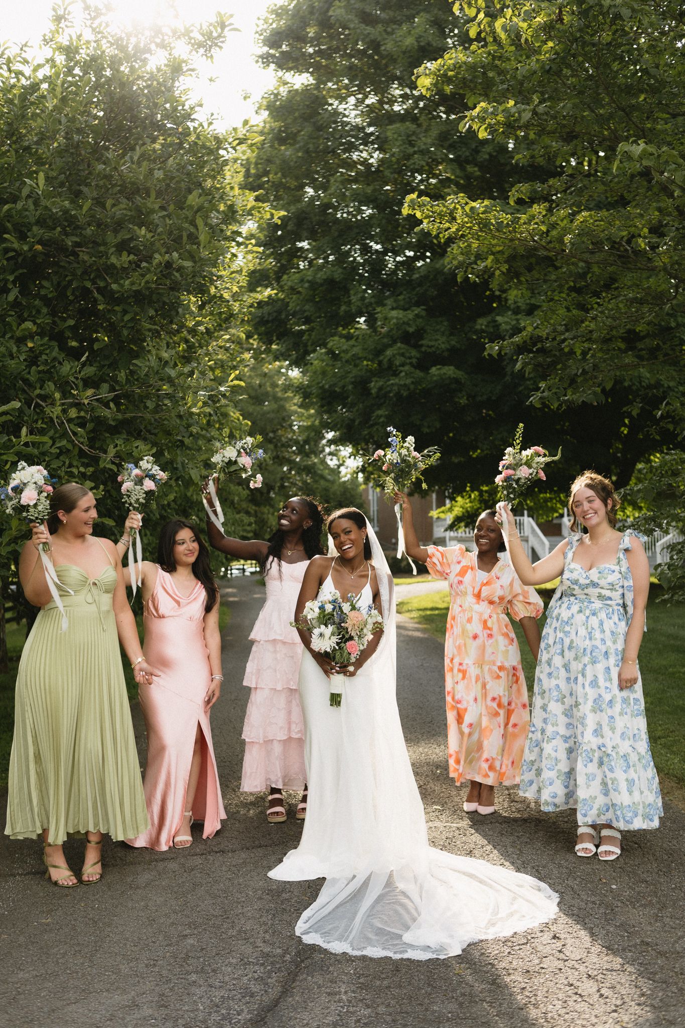 Bridesmaids in colorful flowing dresses celebrate with the bride on a sunlit garden path.