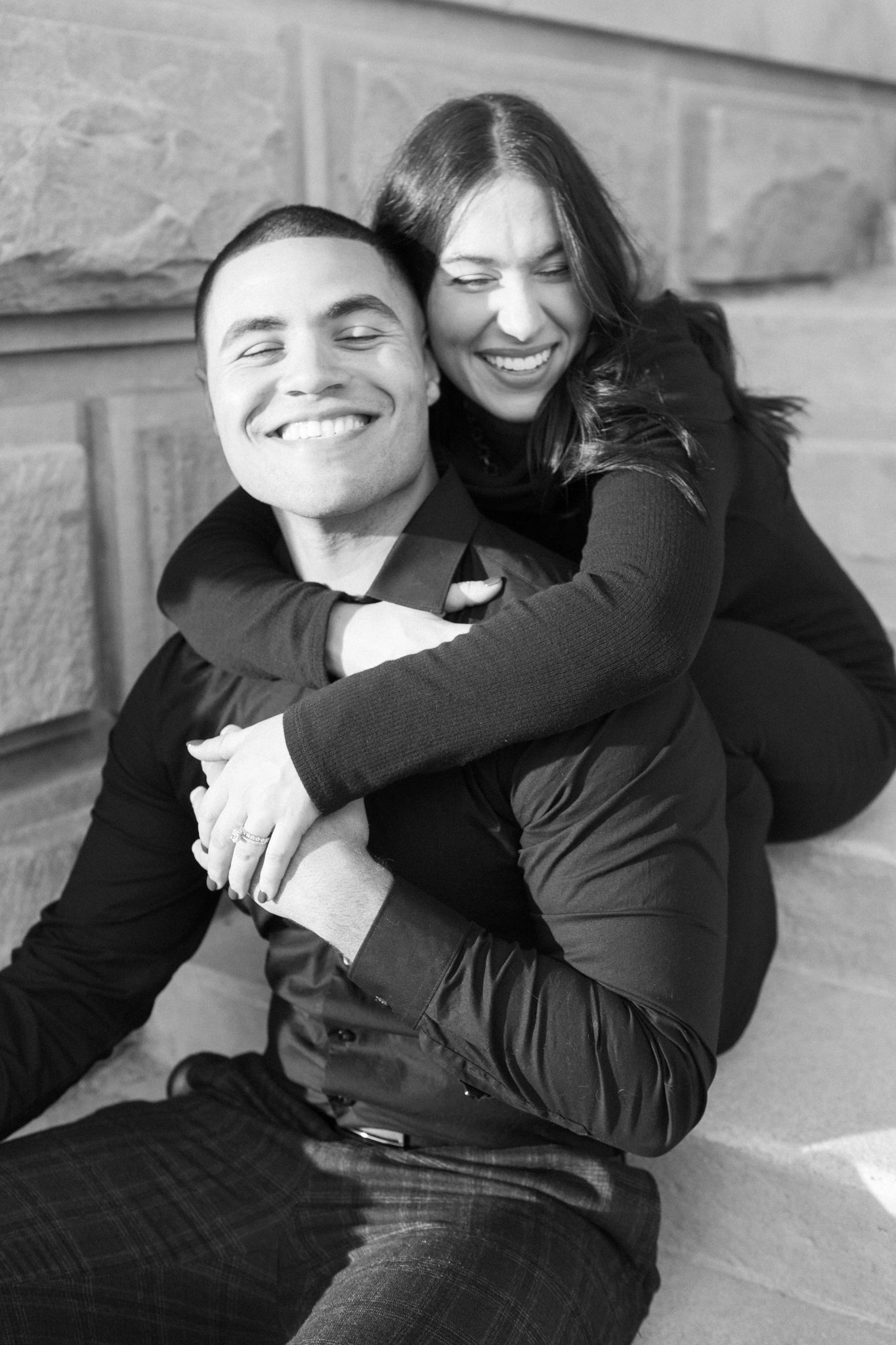 A happy couple embraces and laughs together in a casual black and white portrait against a stone wall.
