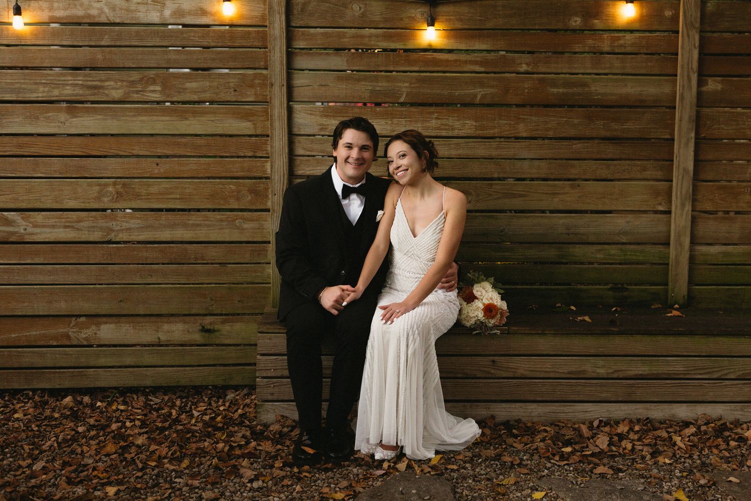 Wedding couple posing against rustic wooden wall with string lights in evening setting.