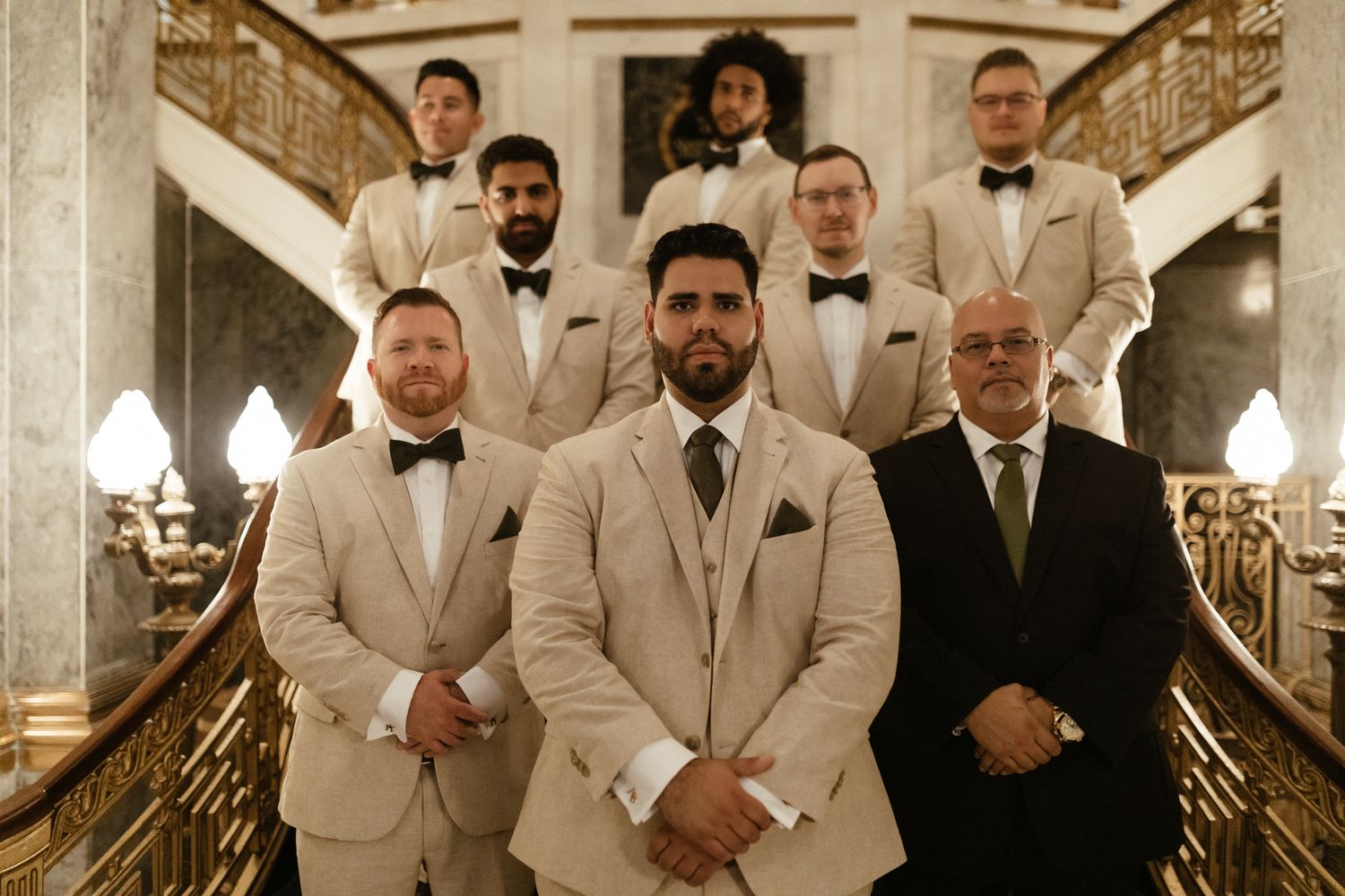 Groomsmen in beige suits and black bow ties stand on an elegant carpeted staircase with decorative railings.
