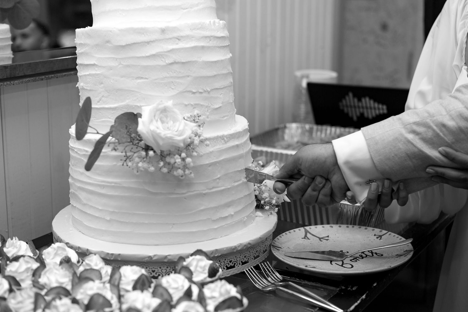 A black and white photo shows a wedding cake being cut at a reception with decorative white frosting layers.