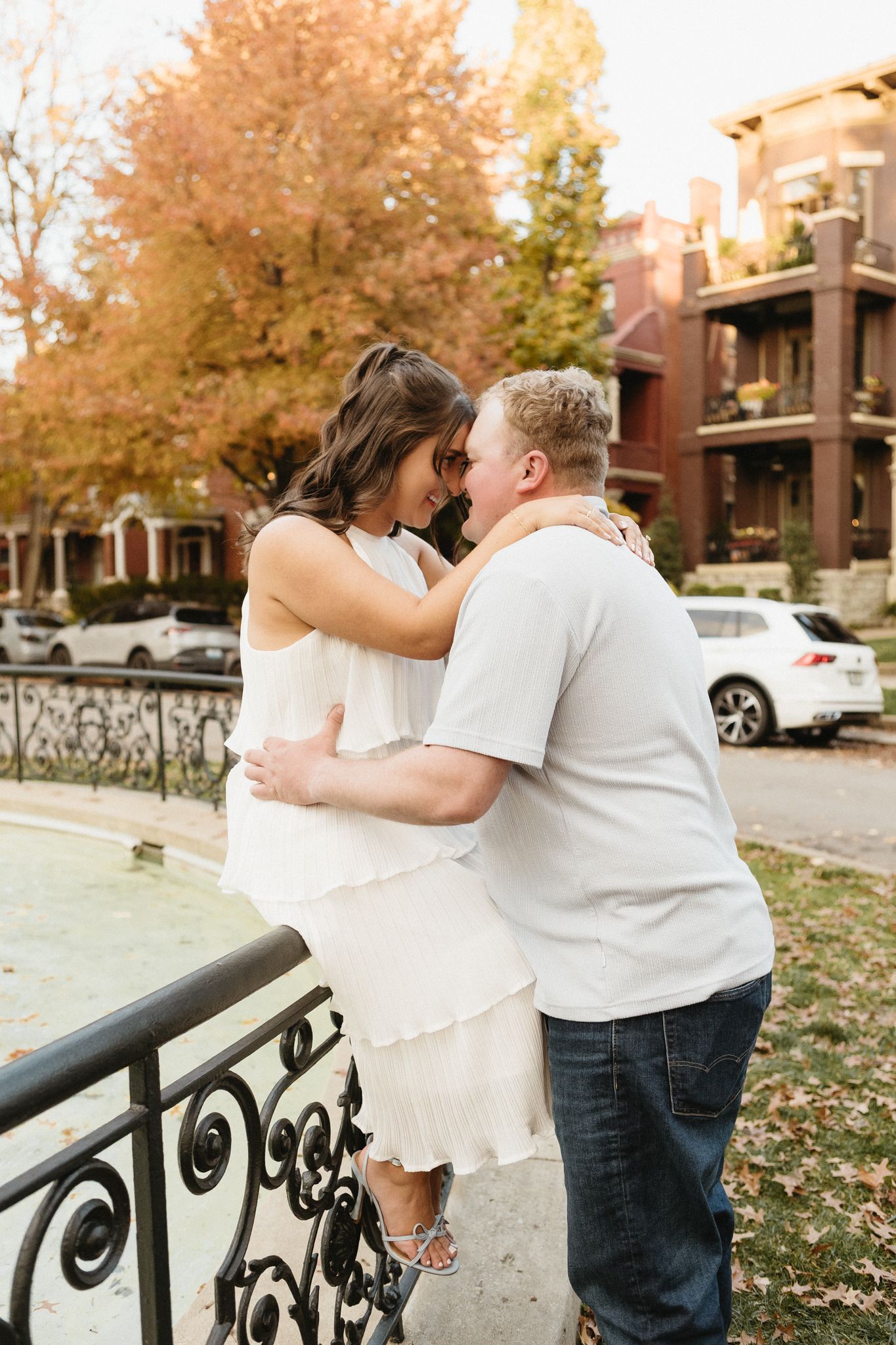A couple embraces near a decorative iron railing during a fall photoshoot with autumn trees in the background.