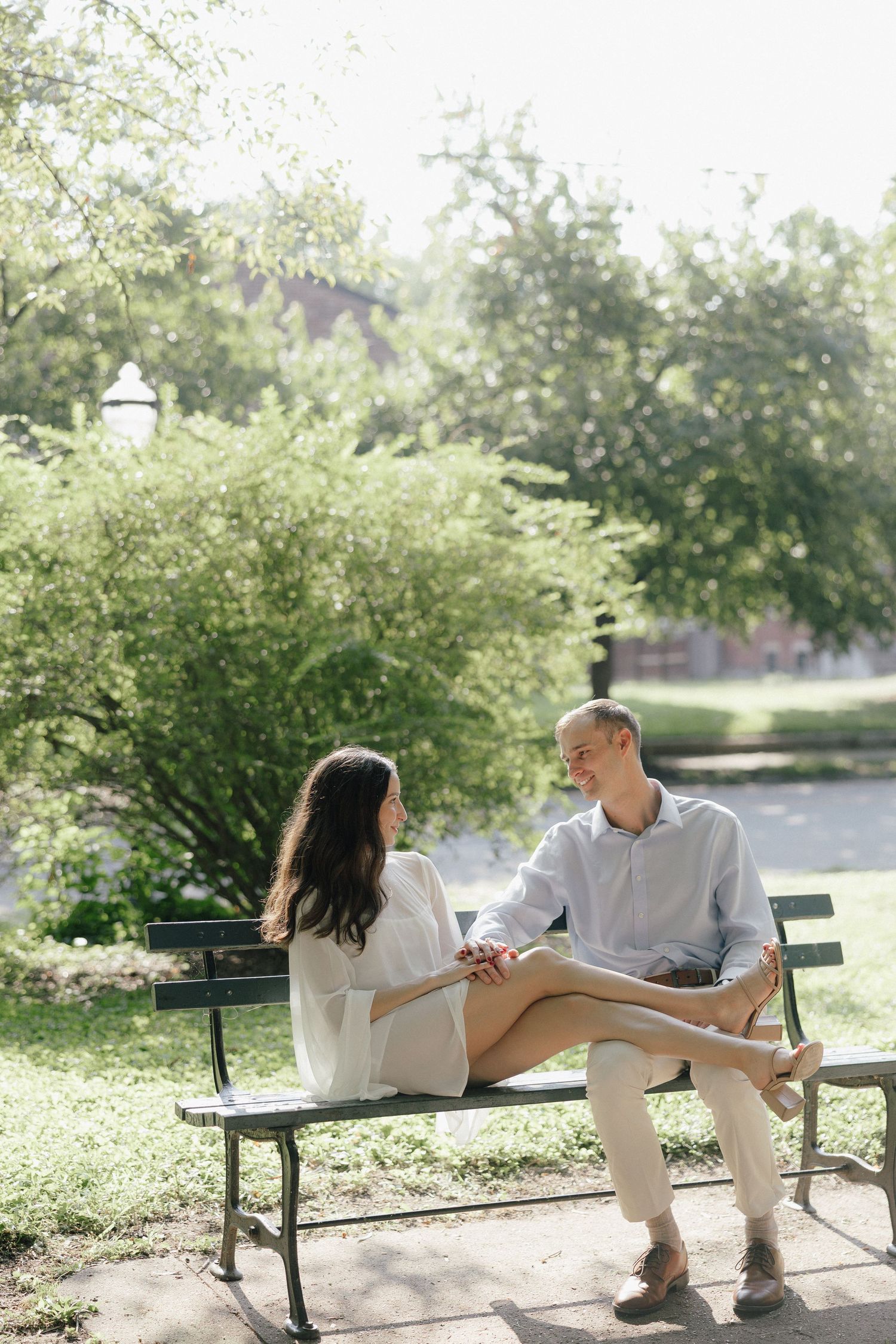 Couple sitting on a park bench holding hands and smiling at each other on a sunny day.