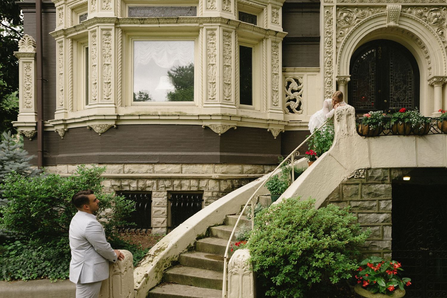 A man in a white suit stands at the base of ornate stone stairs leading to an elegant Victorian mansion.