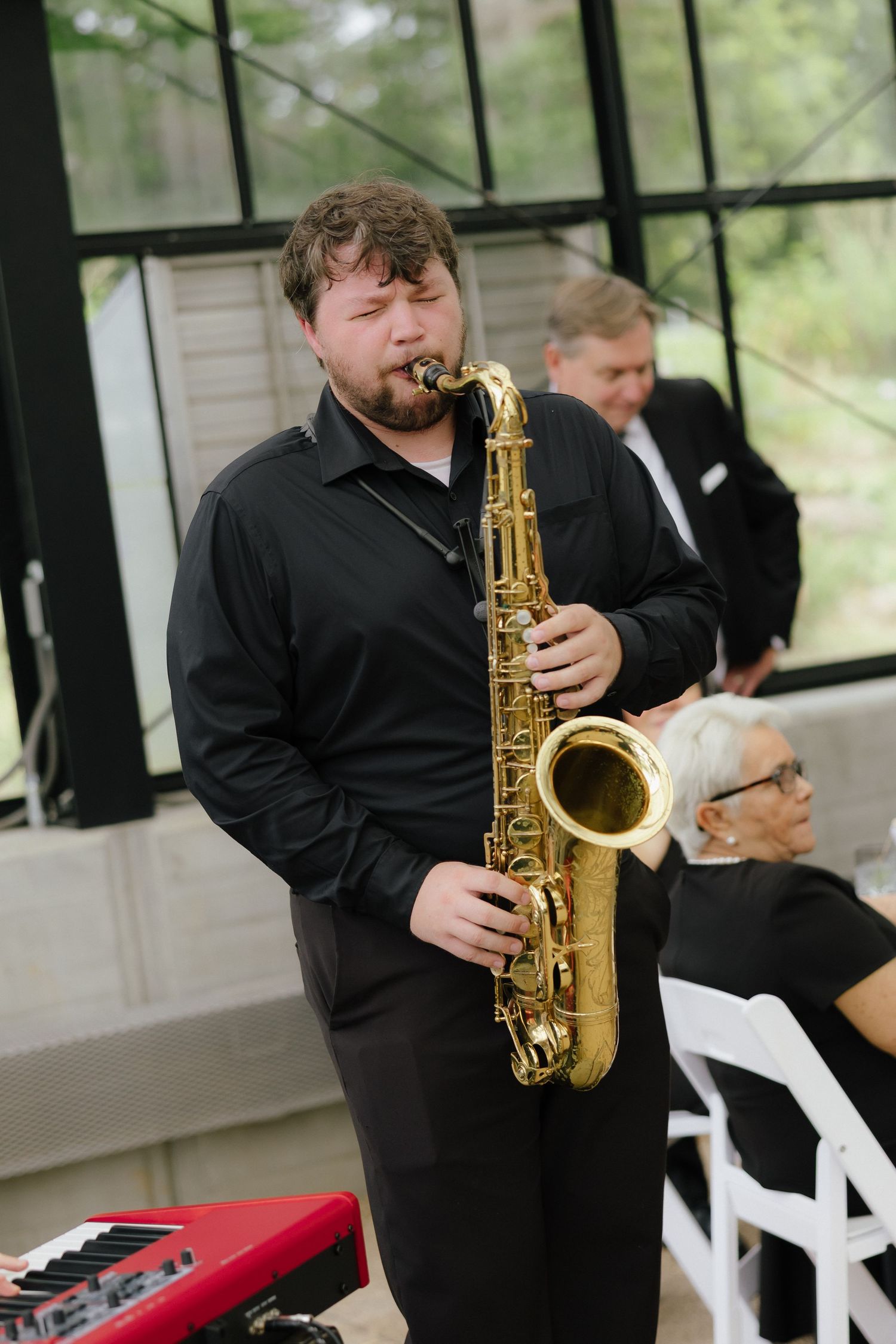 Musician in black shirt plays saxophone at outdoor wedding reception with guests seated nearby.