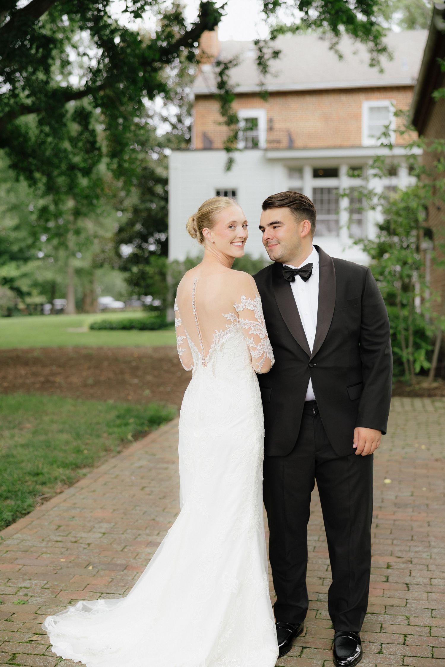 Bride in lace long-sleeve wedding gown smiling over shoulder with groom in black tuxedo on brick path.