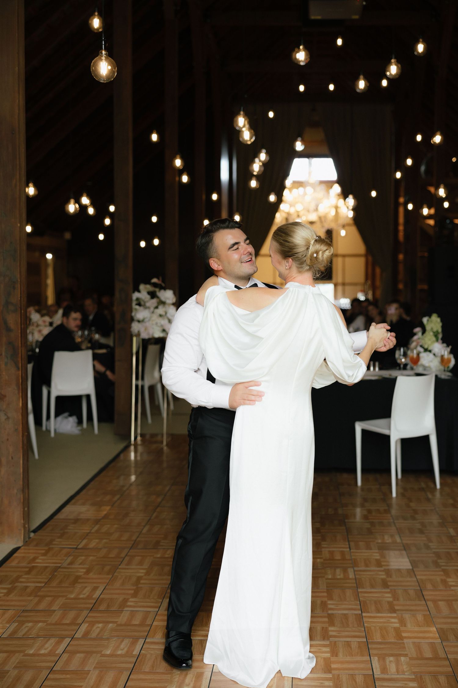 Bride and groom share their first dance under warm string lights in an elegant barn wedding venue.