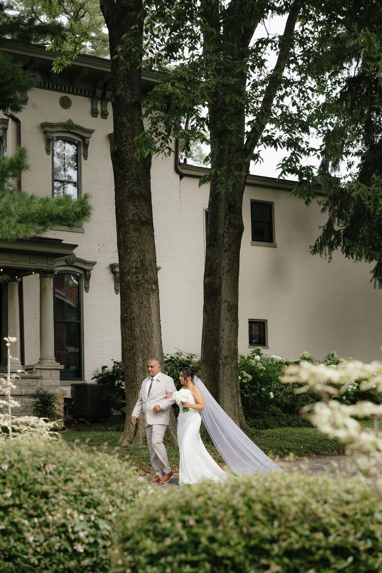 Bride and groom walk hand in hand through lush garden grounds beside a elegant white historic mansion.