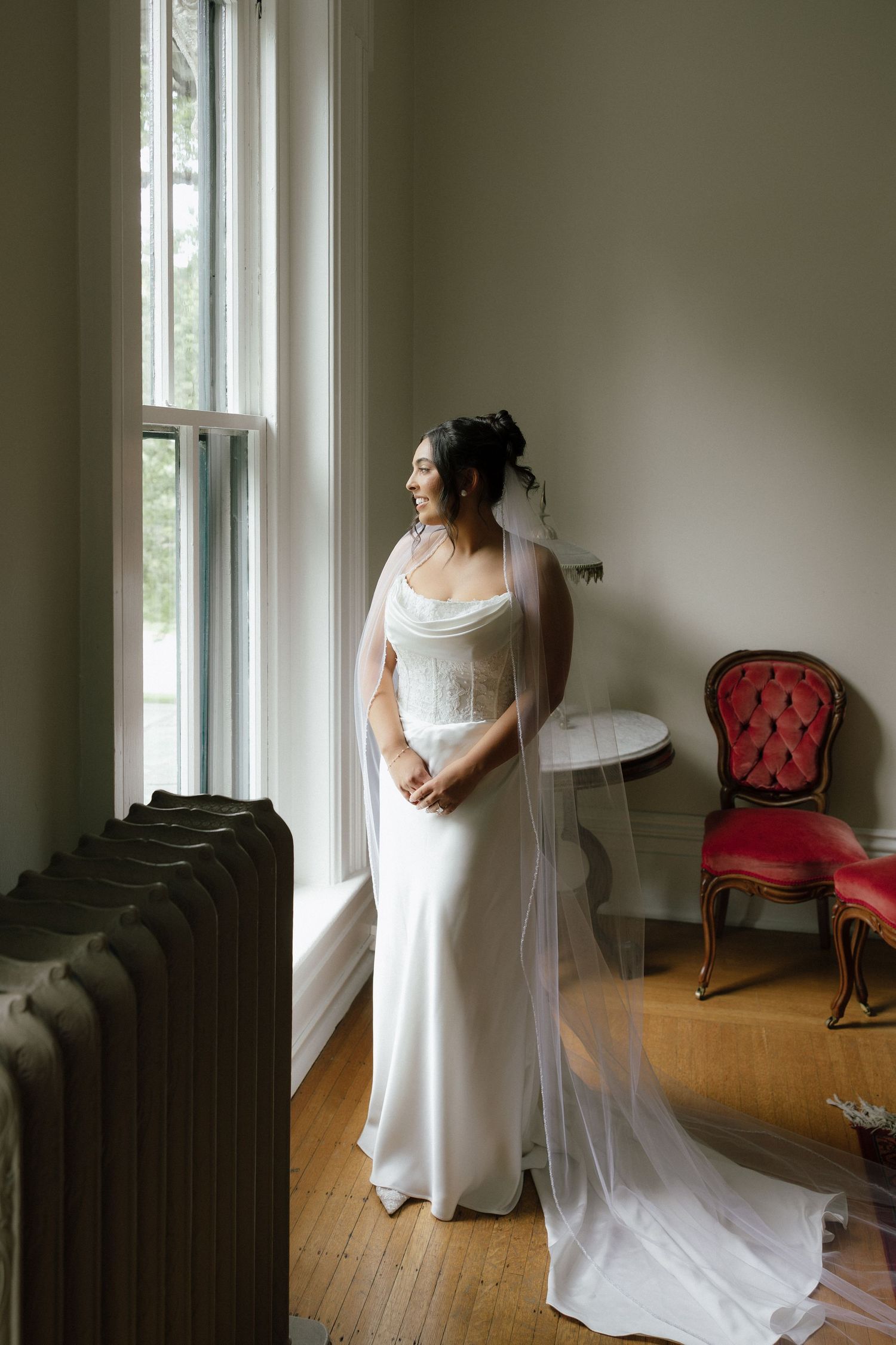 Elegant bride in white cowl neck gown and long veil gazing out tall window on wedding day.
