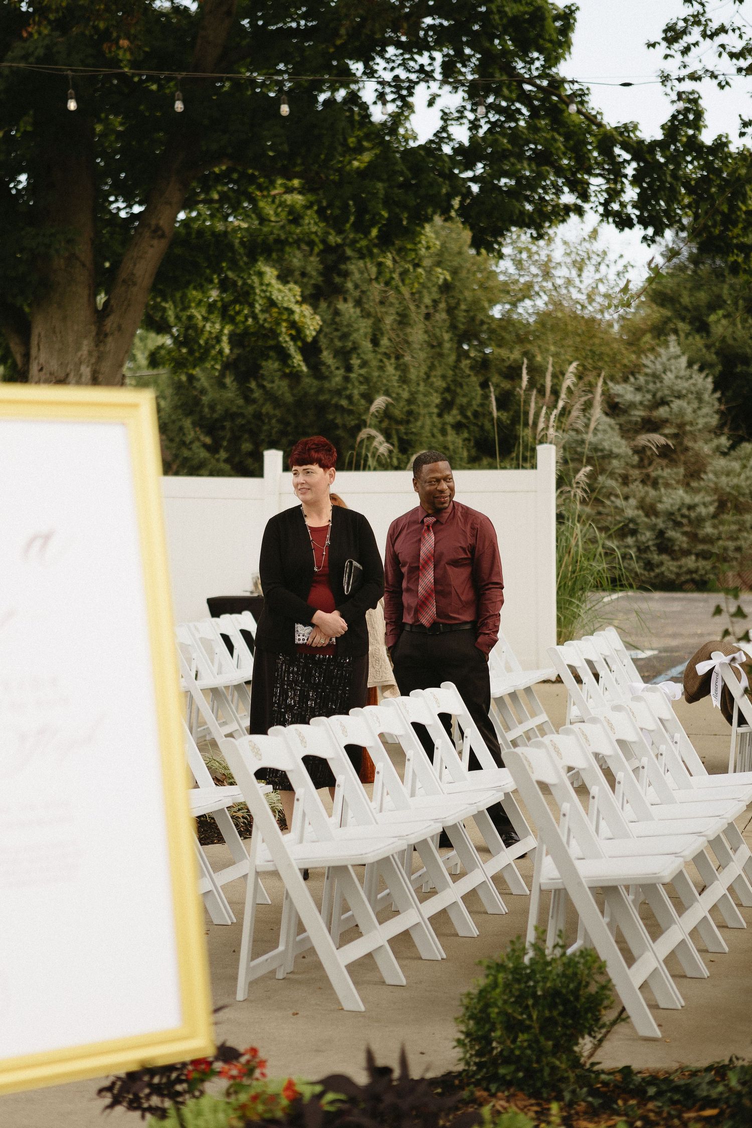 Wedding ceremony setup outdoors with rows of white folding chairs and trees in the background.