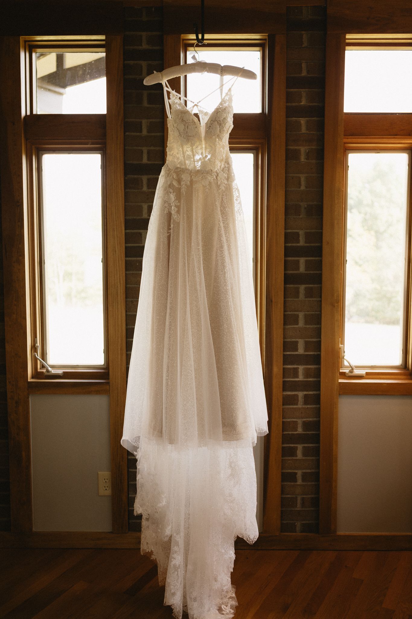 A white wedding dress hangs in a window with natural light streaming through, creating a romantic silhouette.