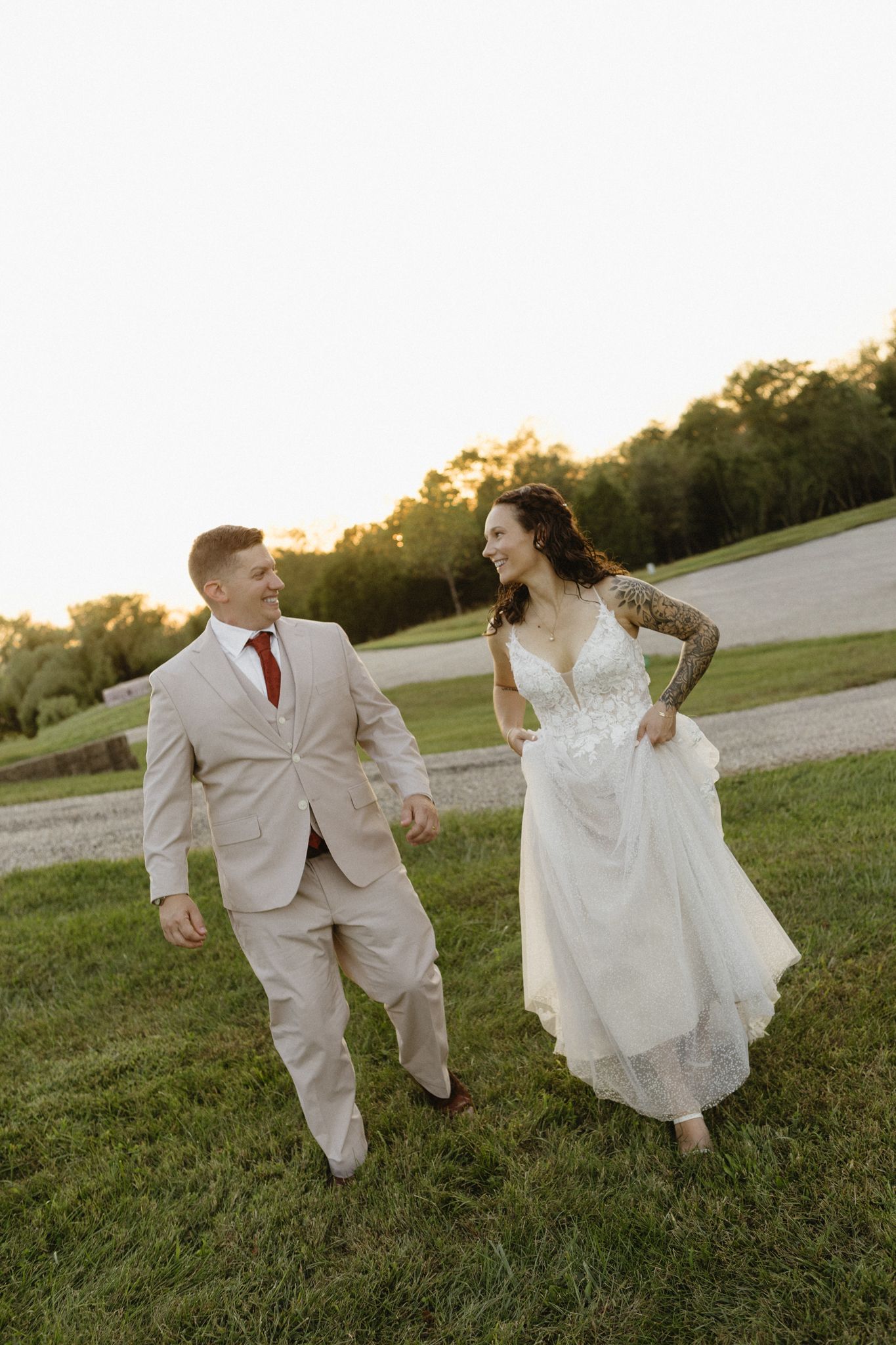 Couple running and dancing together on a grassy field at sunset in wedding attire.