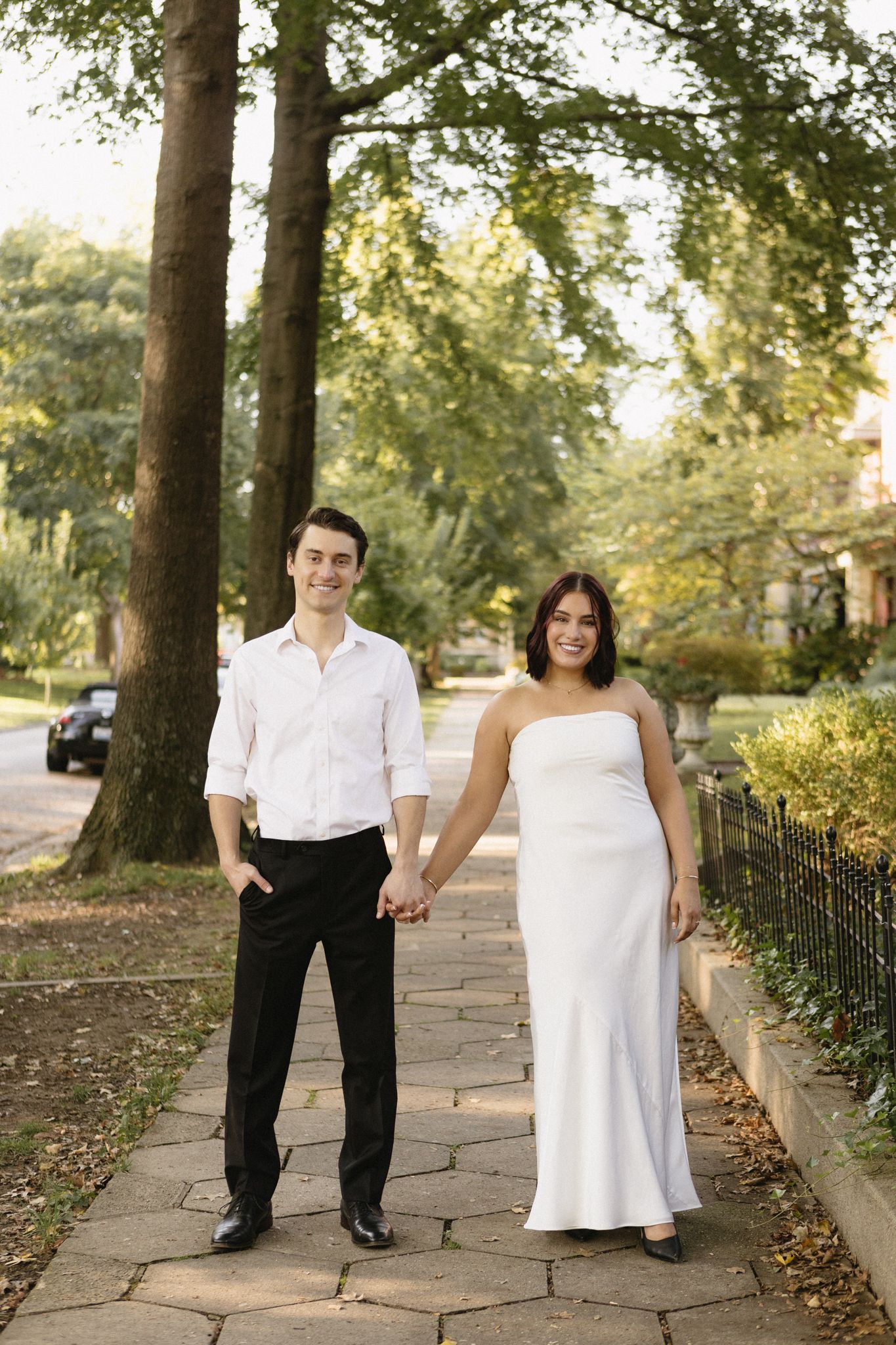 A couple in formal attire poses together on a stone pathway surrounded by trees in a park setting.