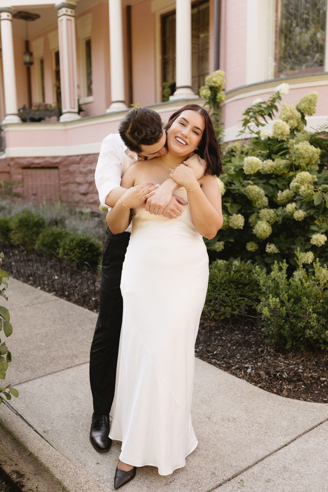 A tender embrace shared between a couple on a walkway with pink columns and landscaping.