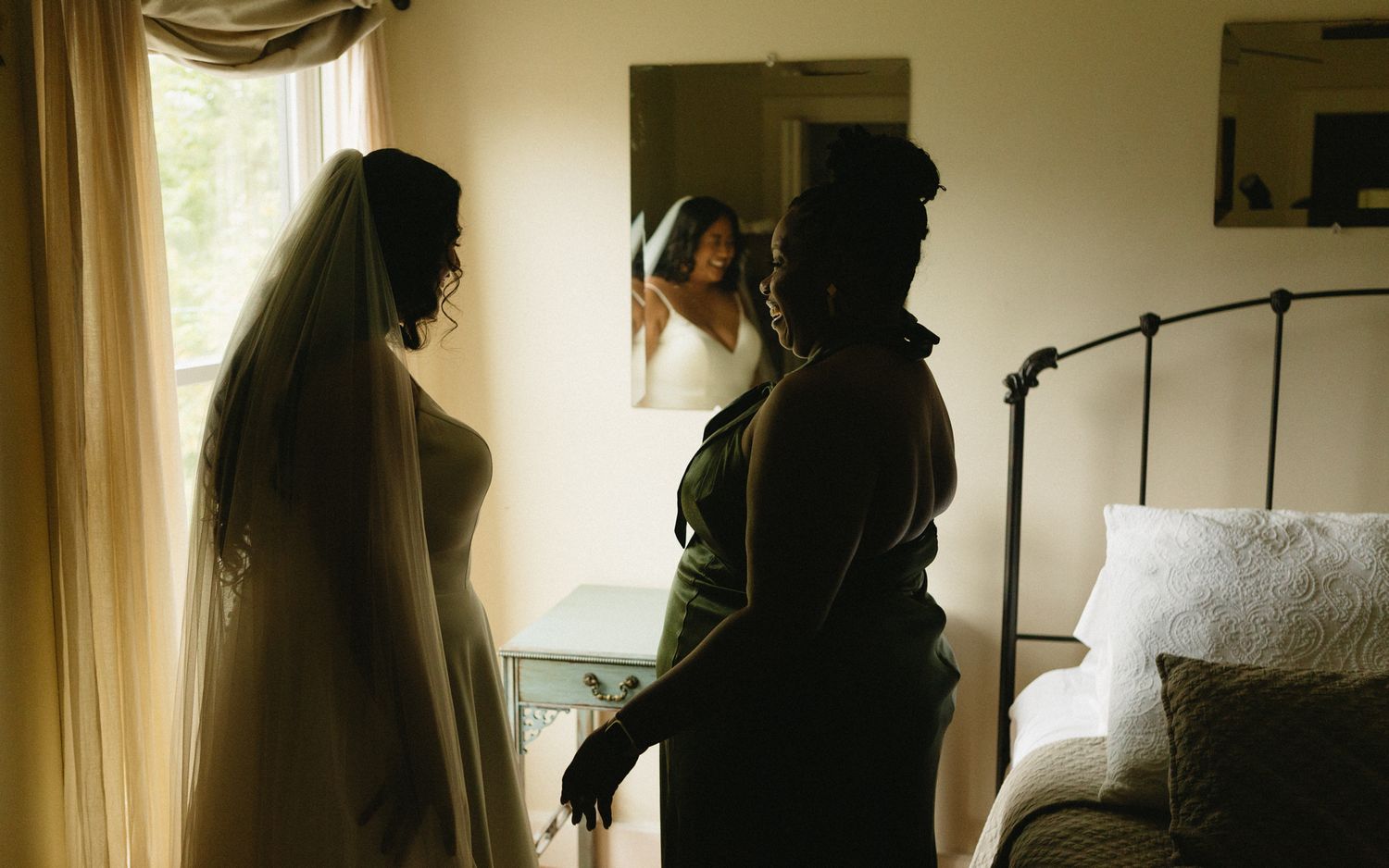Two figures in wedding attire getting ready in a sunlit bedroom with a vintage wrought iron bed frame.