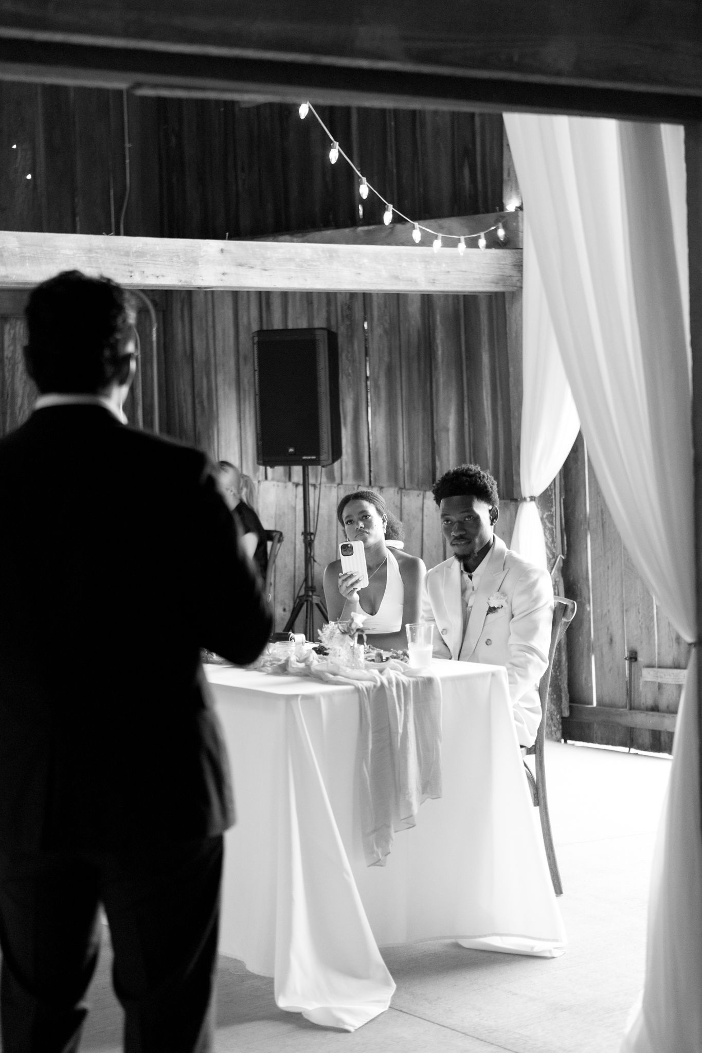 Black and white photo showing an elegant reception venue with draped white curtains and bistro lights.