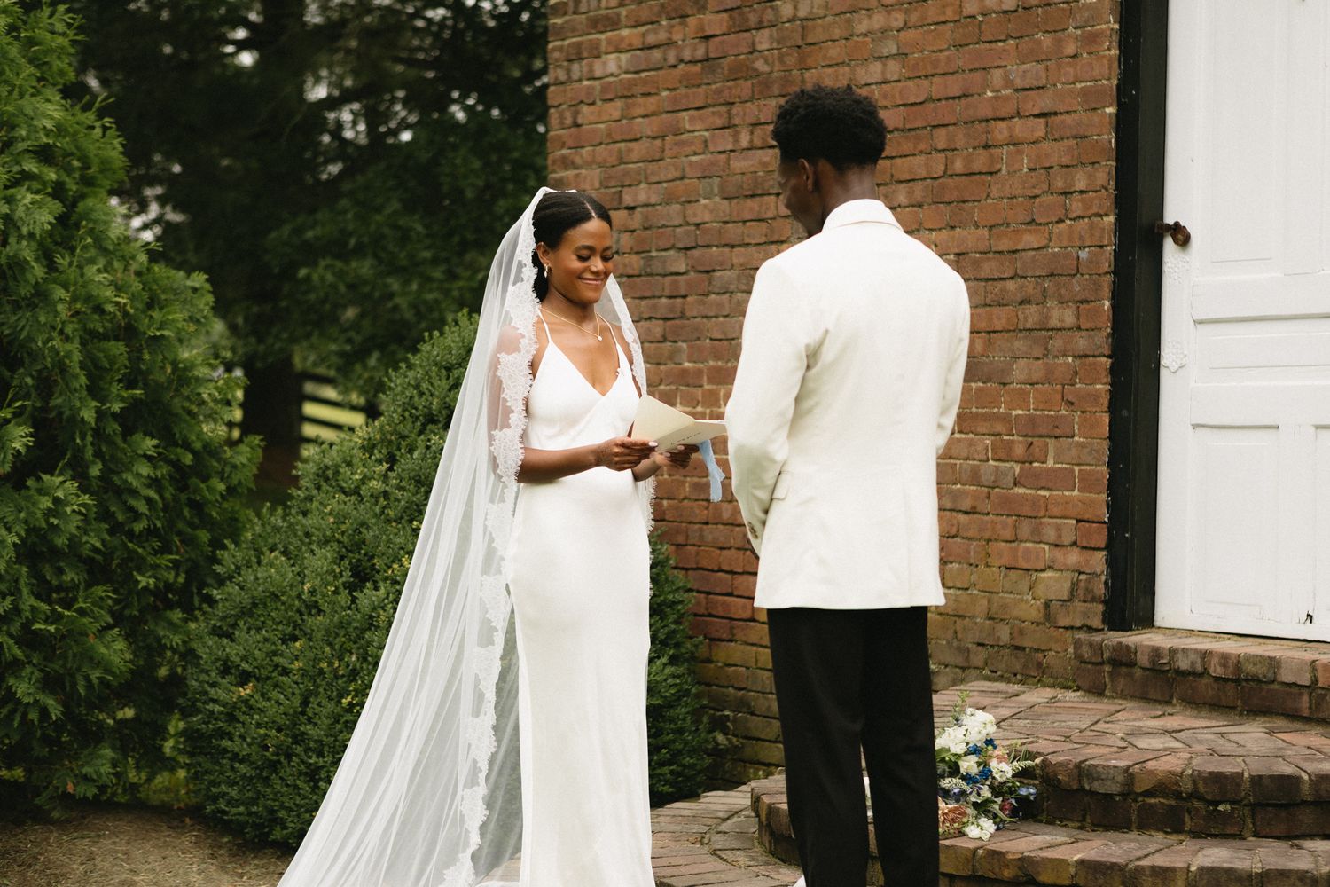 Romantic wedding couple poses against brick wall exterior with lush greenery.