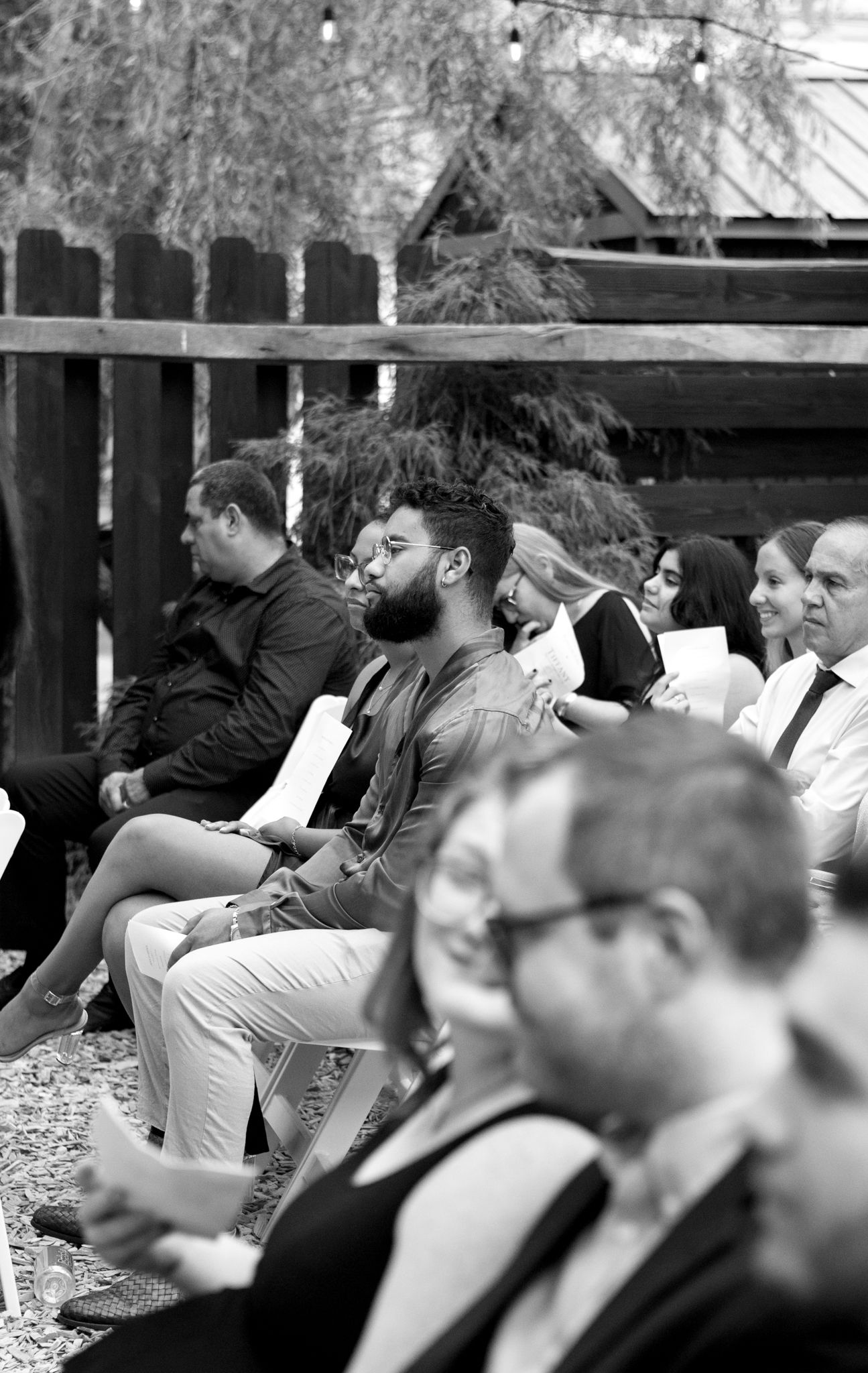 A black and white photo showing a group of people seated outdoors in front of a wooden fence during an event.