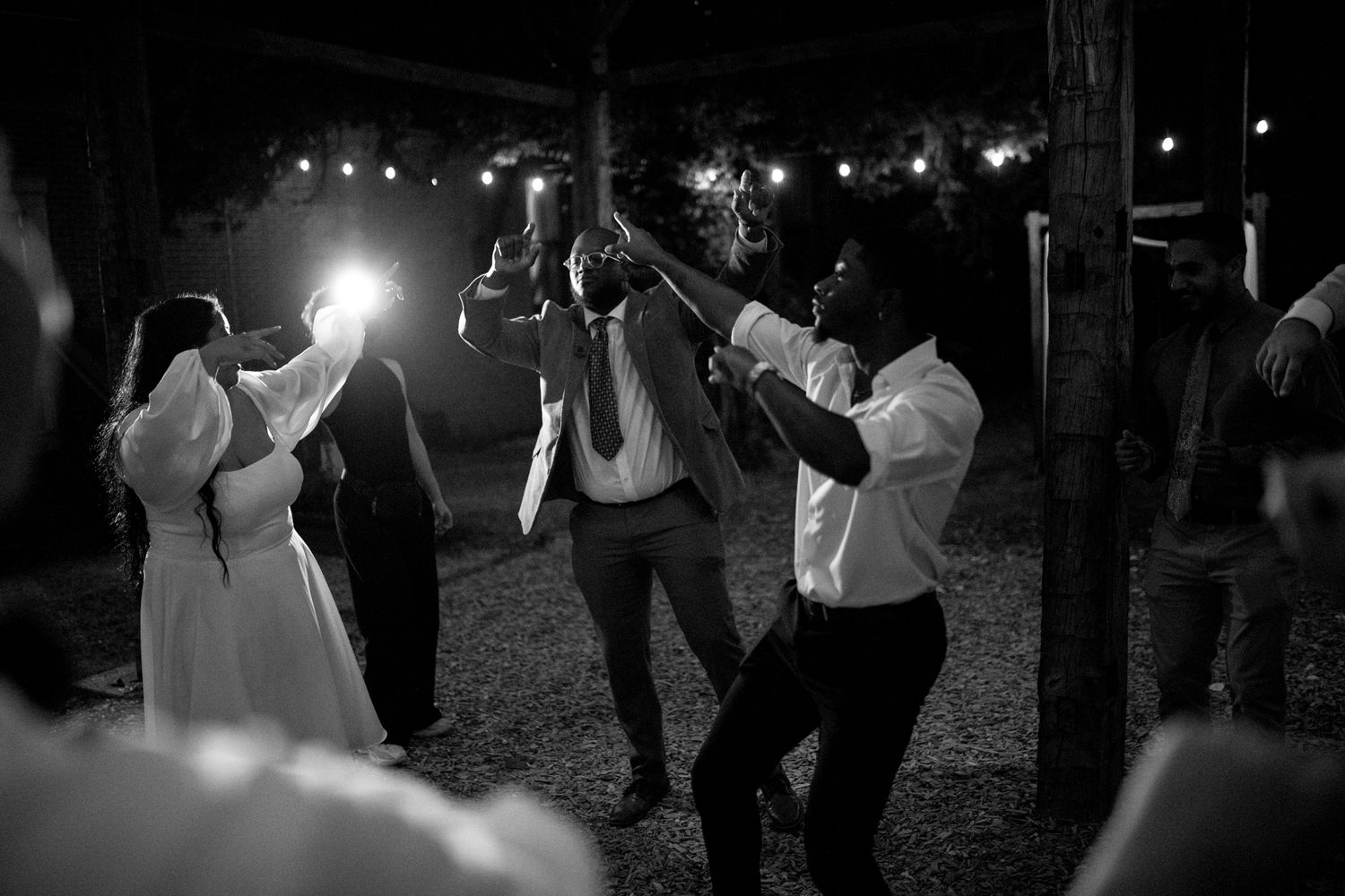 A group of people dancing and celebrating at night under string lights at an outdoor party.