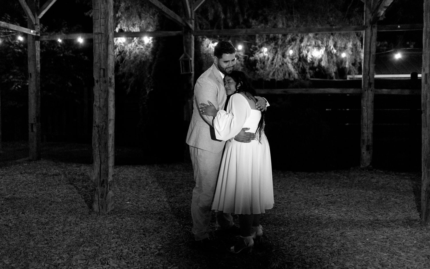 A couple embraces during an outdoor evening reception under twinkling lights strung between trees.