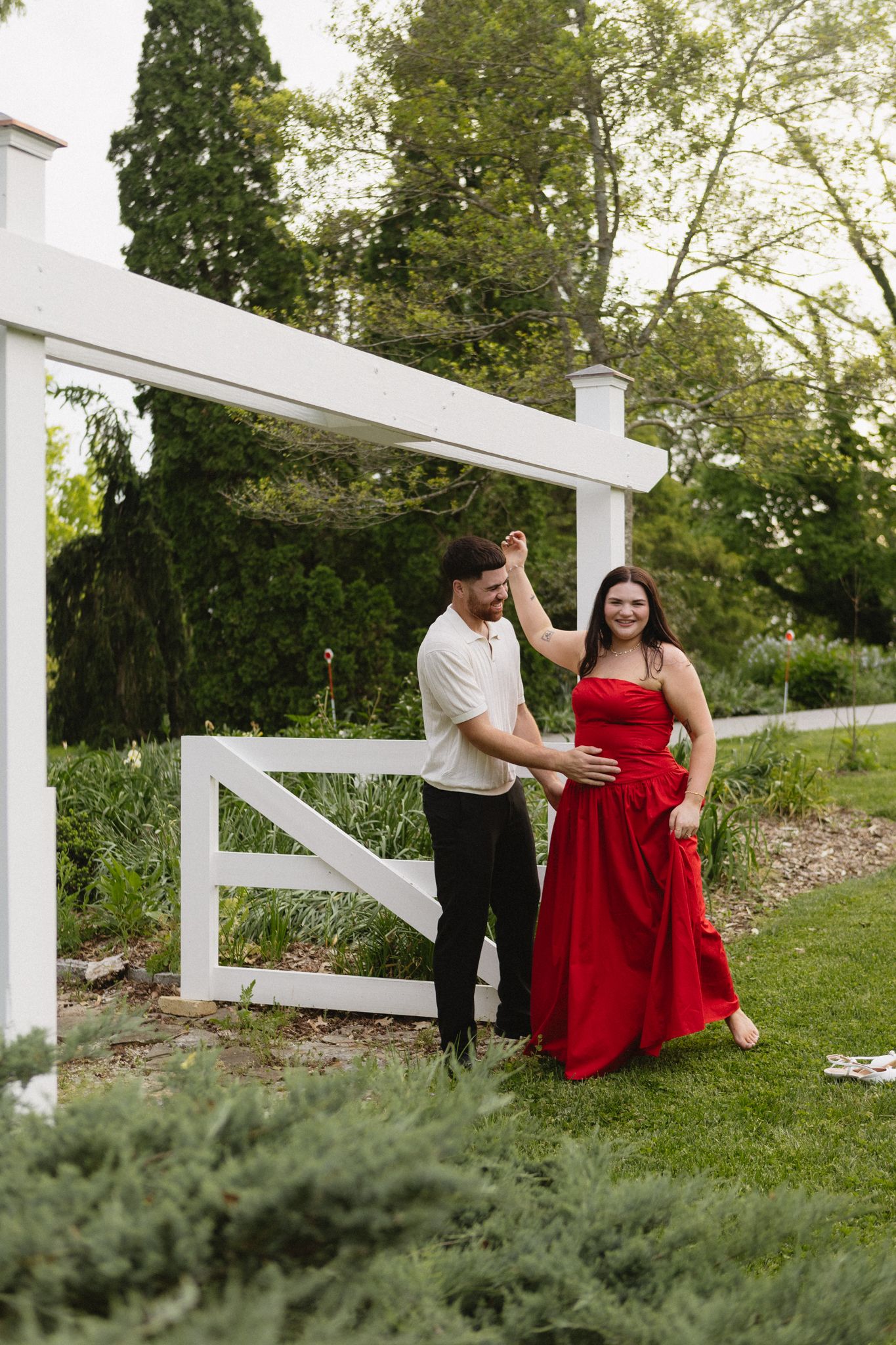 Couple poses playfully by white fence in garden setting with beautiful greenery.