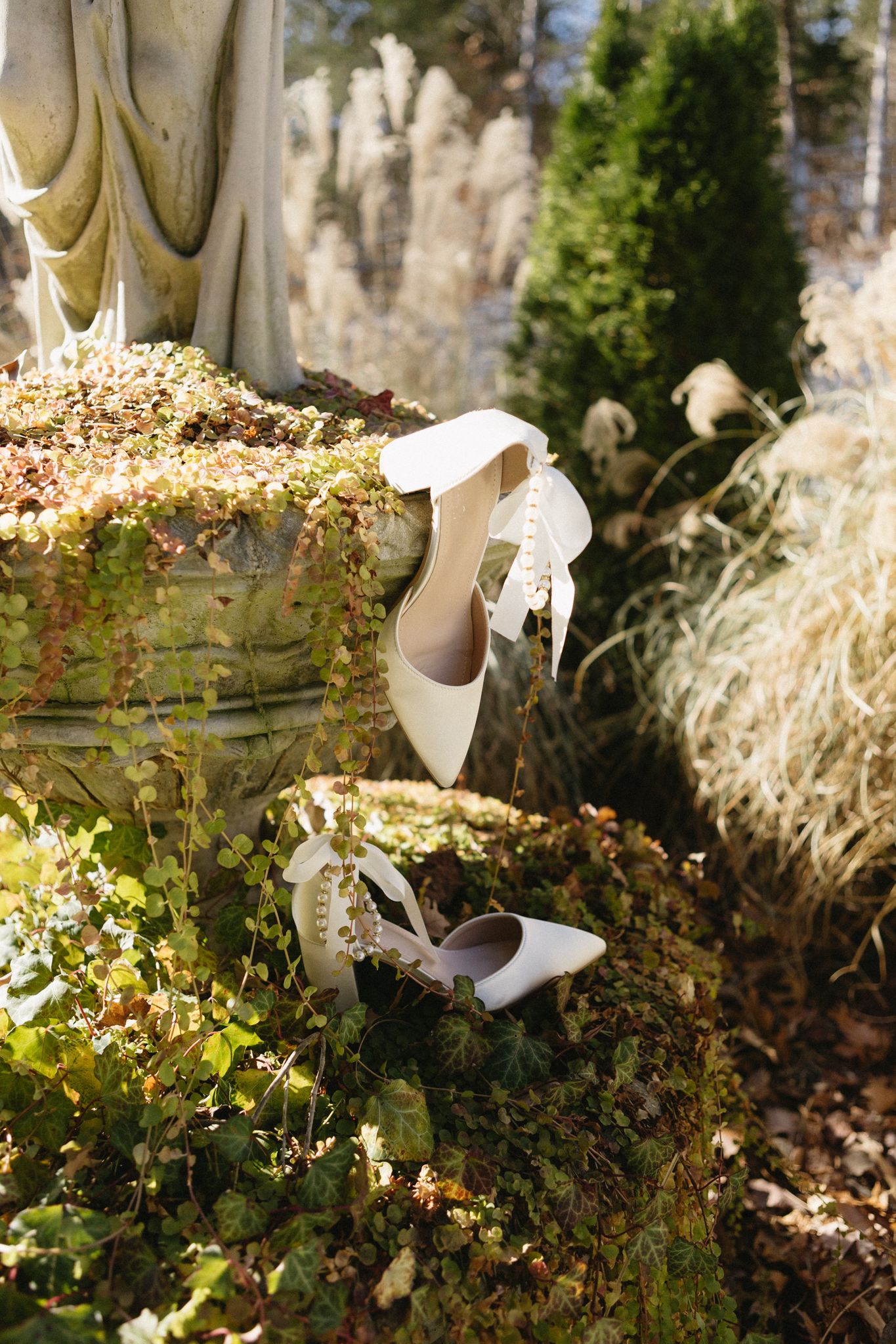 Elegant white bridal shoes with lace details displayed on a mossy garden urn in natural outdoor setting.