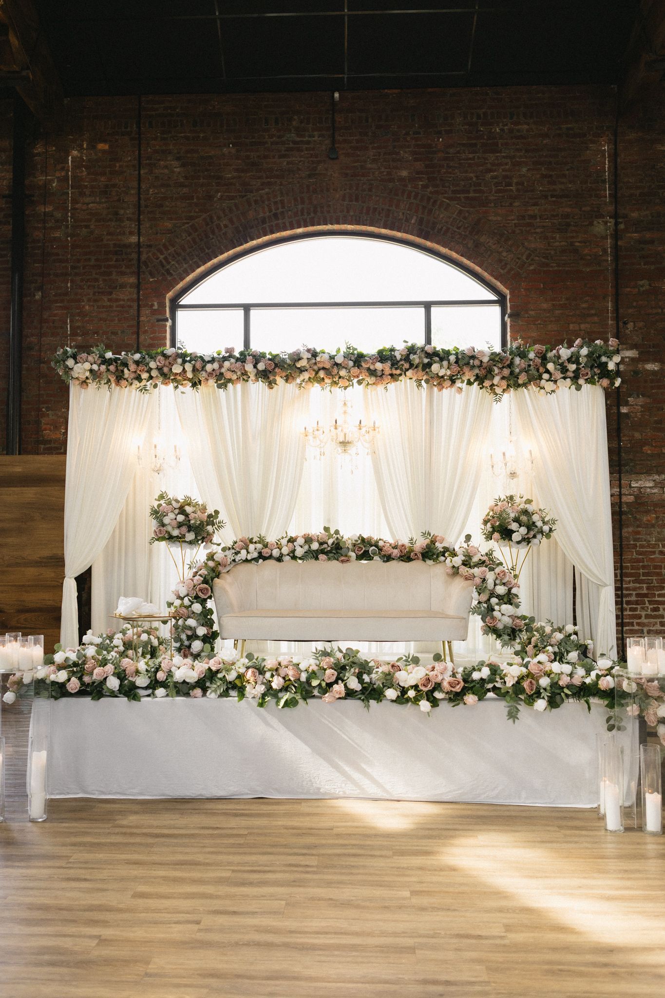 Wedding ceremony backdrop with white draping and floral arrangements against exposed brick wall in industrial venue.