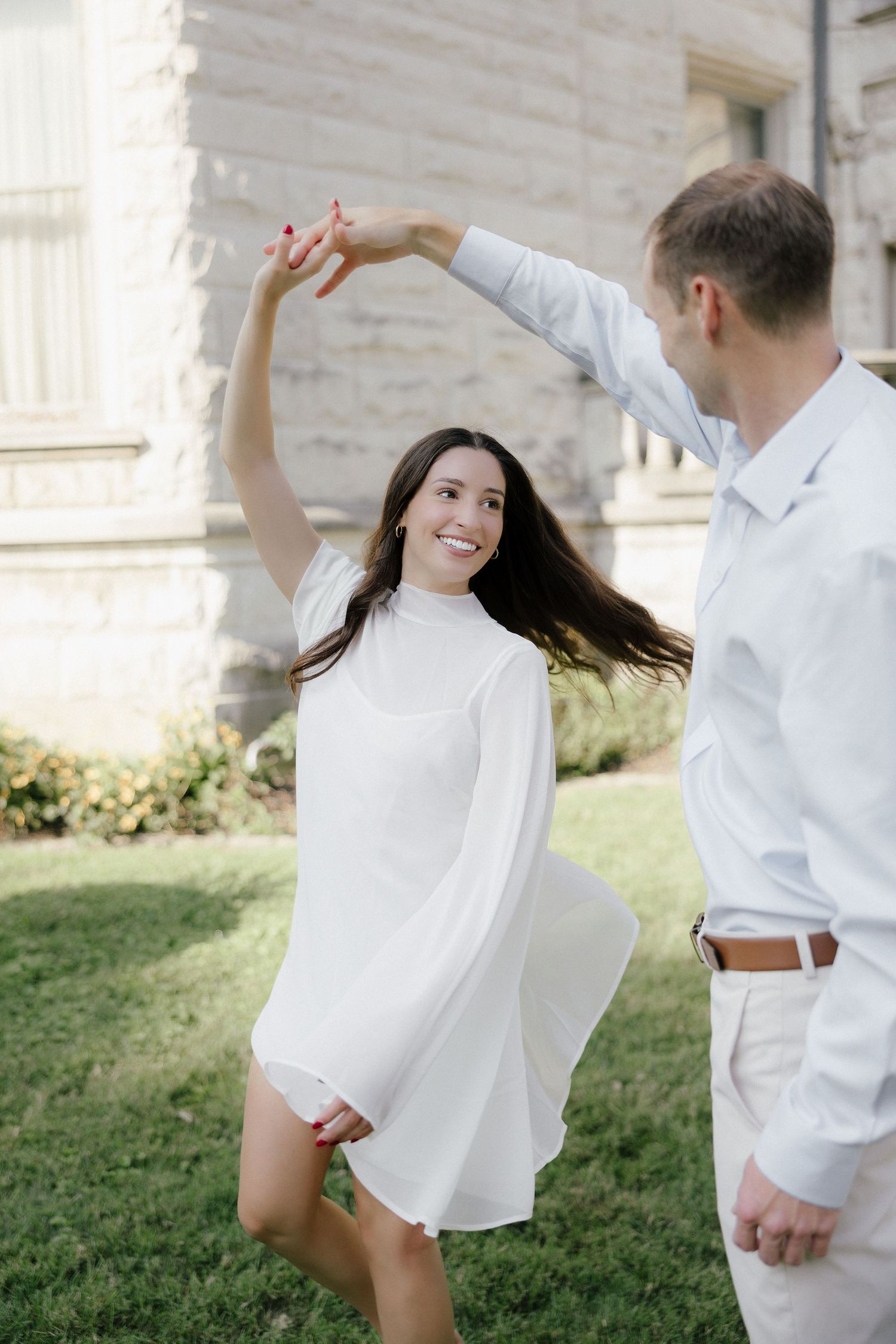 Joyful couple dancing outdoors, woman in white dress spinning gracefully in front of a stone building.