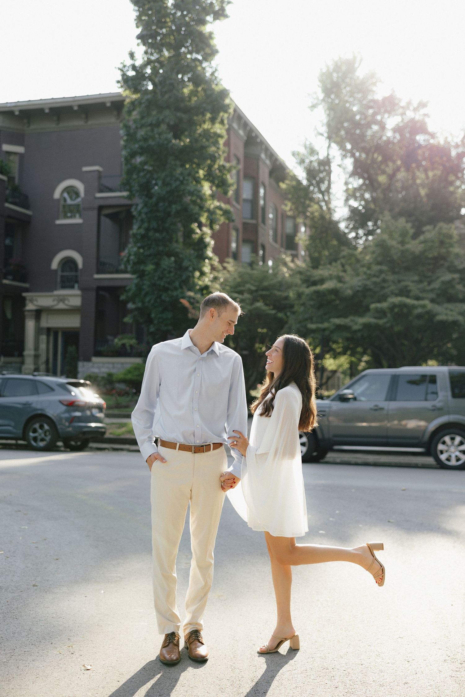 Engaged couple holding hands on a sunny street, woman in white dress lifting one foot playfully.