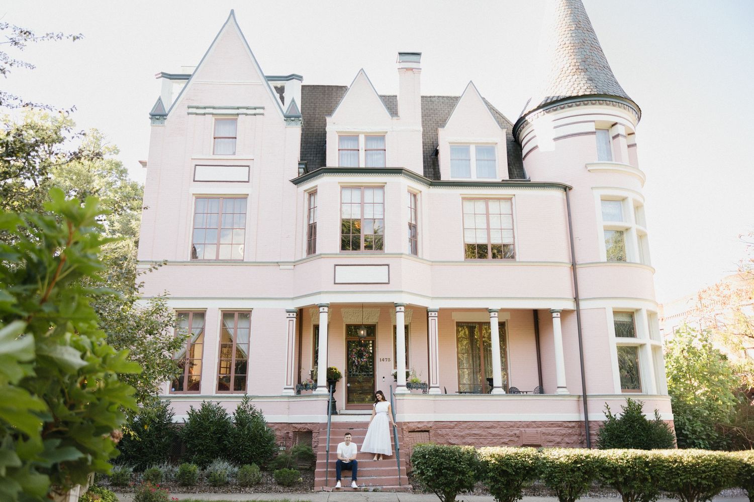 Elegant pink Victorian mansion wedding venue with turret, white columns, and bride standing at entrance surrounded by lush greenery.