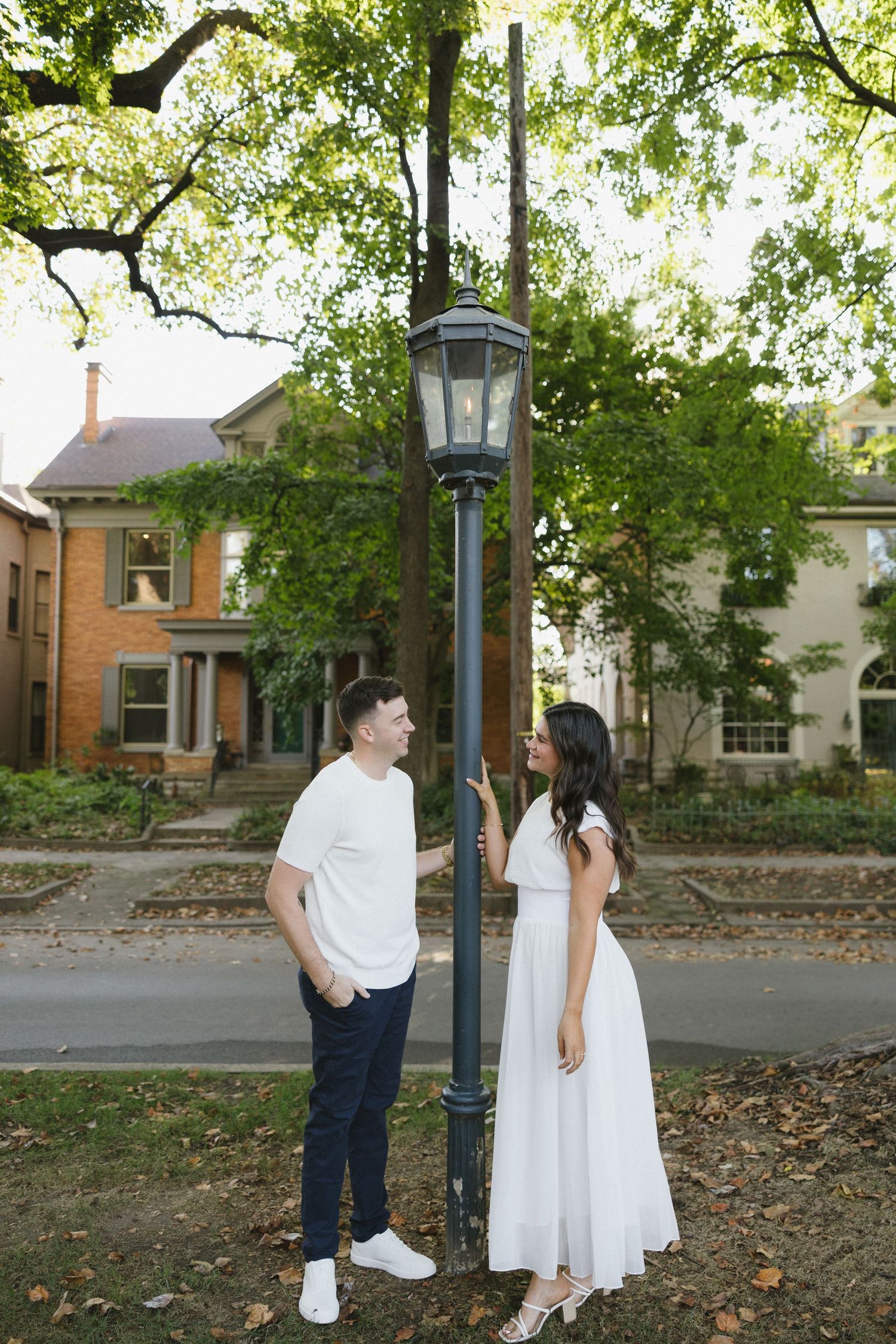 A couple stands by a vintage street lamp surrounded by lush trees and historic homes, the woman in a white dress smiling at her partner.