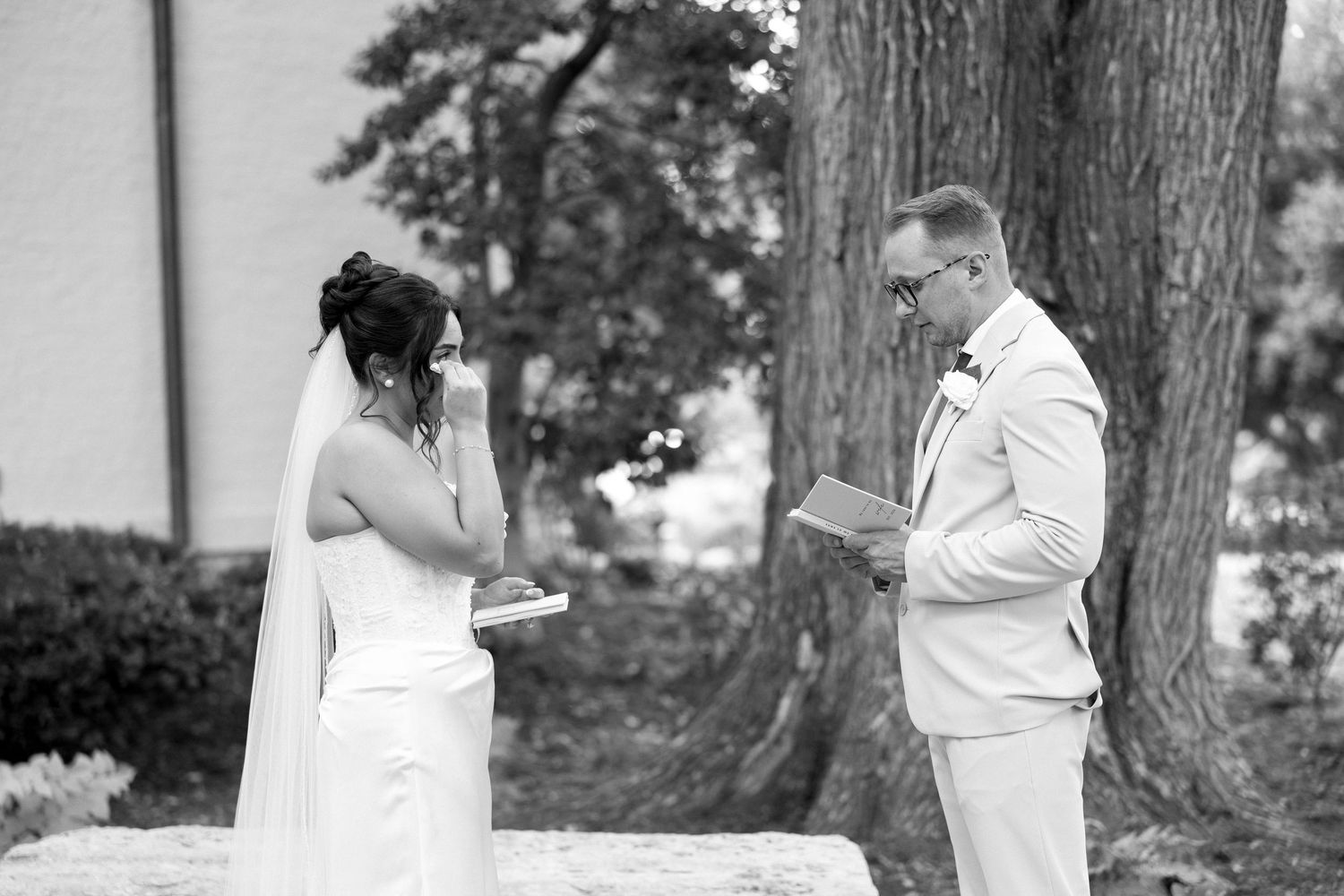 Black and white wedding photo of bride wiping tears while groom reads vows outdoors near a large tree.