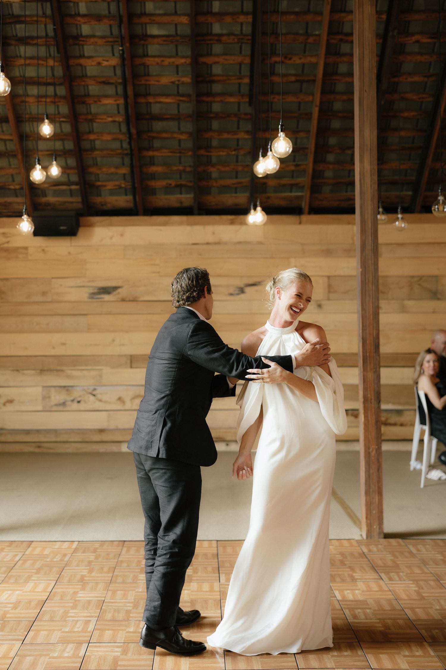Bride in white off-shoulder gown and groom share first dance in rustic barn venue with Edison lights.