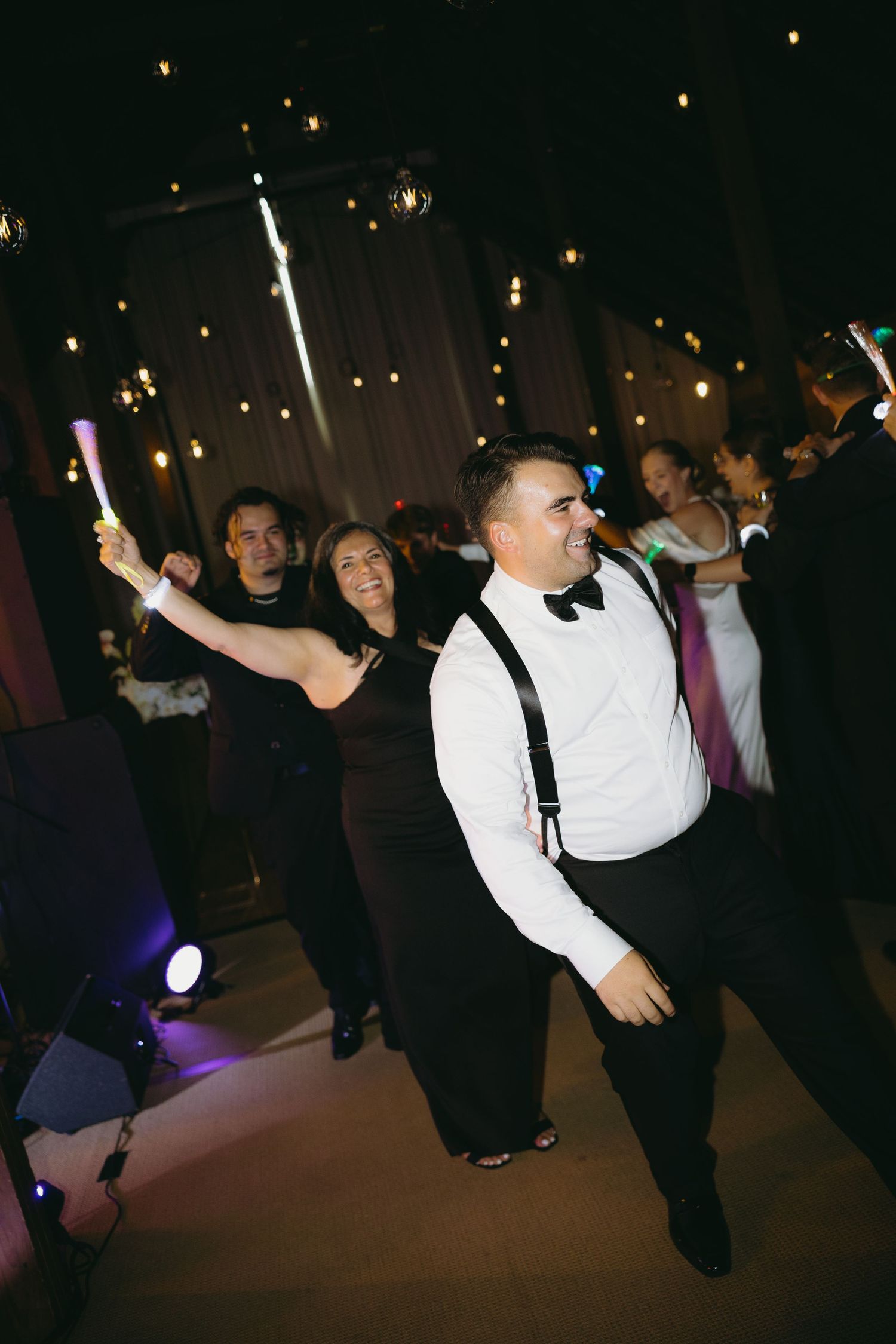Groom in white shirt and suspenders dances joyfully with guests holding glow sticks at reception.