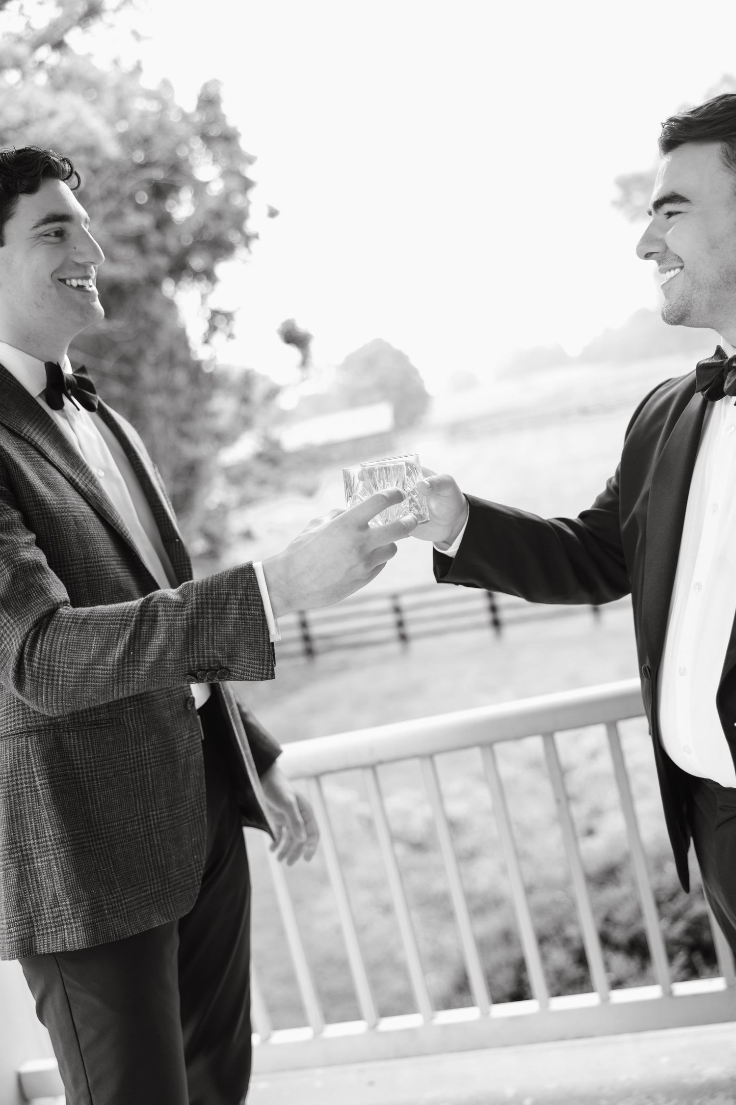 Black and white photo of two men in tuxedos toasting drinks outdoors at wedding celebration.