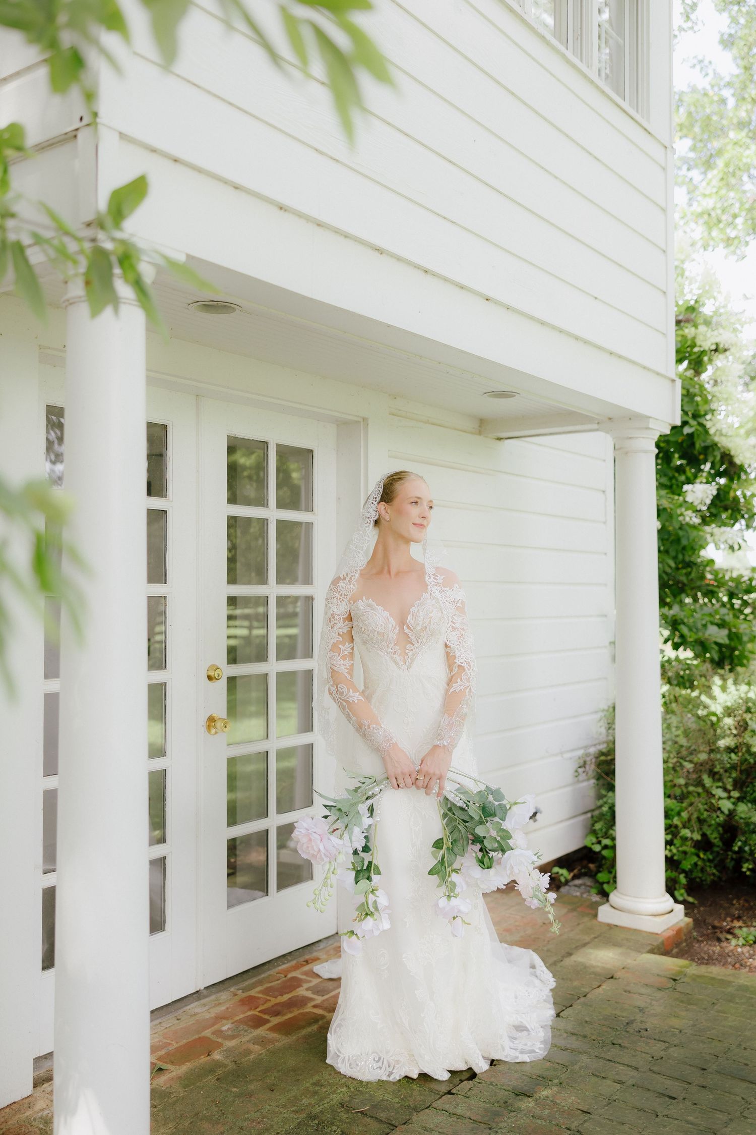 Bride in long-sleeve lace mermaid wedding gown holding floral bouquet outside white colonial building.