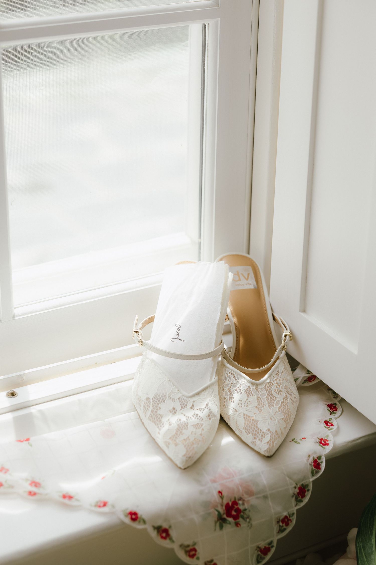 Elegant white lace bridal heels with ankle straps displayed on a windowsill with a floral handkerchief.
