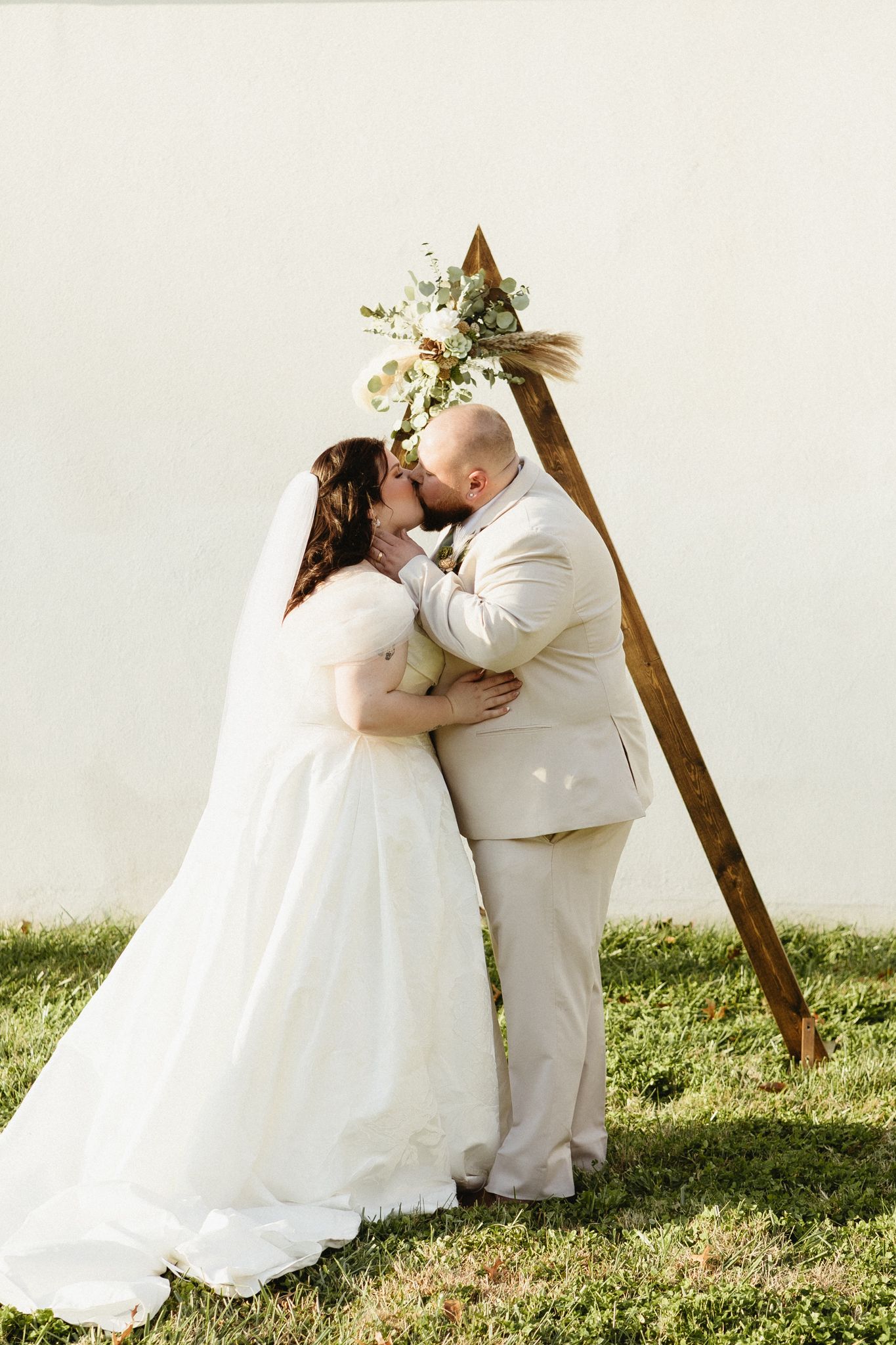 A romantic outdoor wedding ceremony under a wooden triangle arch decorated with white flowers and greenery.