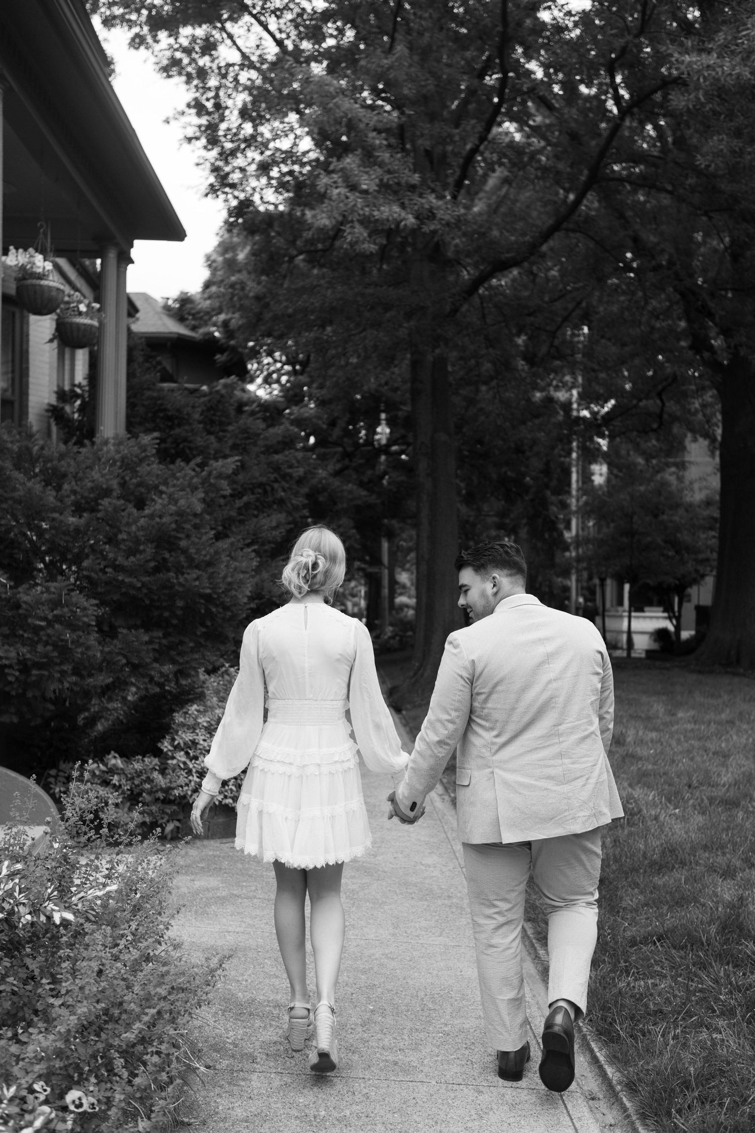 Black and white photo of a couple holding hands walking down a tree-lined sidewalk, woman in white dress, man in light suit.