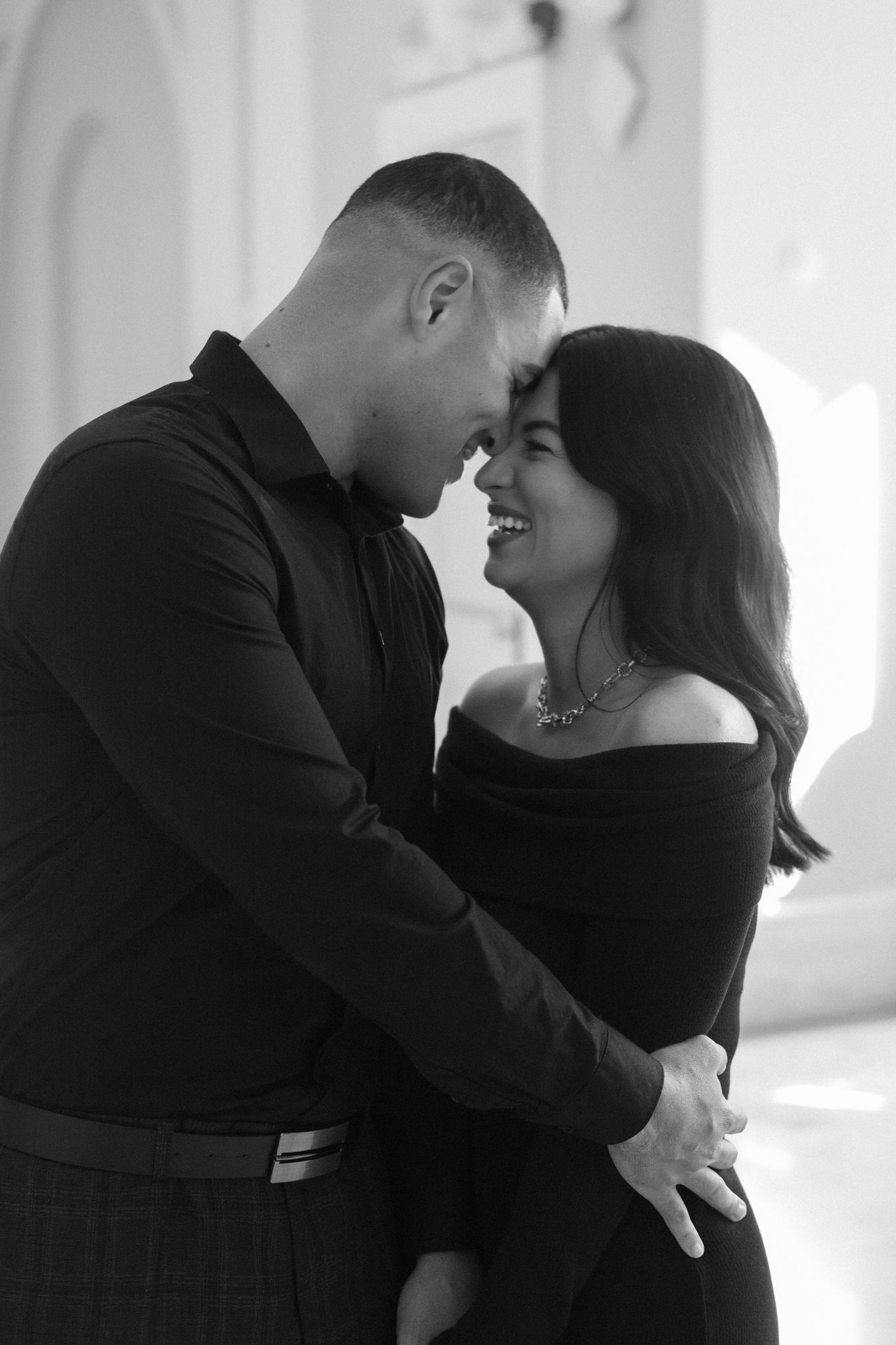 Romantic black and white portrait of a couple in formal wear sharing an intimate moment in a bright hallway.
