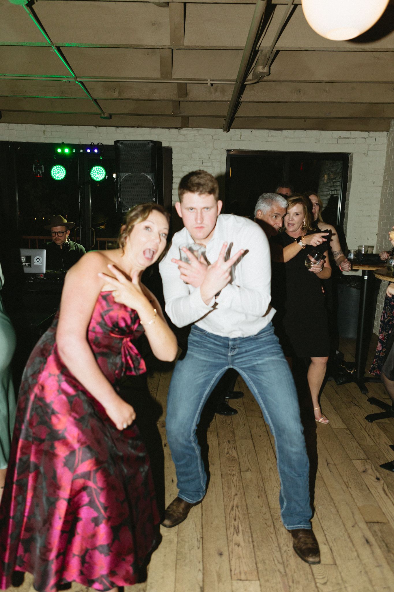 Guests dancing enthusiastically at an indoor party with stage lights and exposed ceiling.